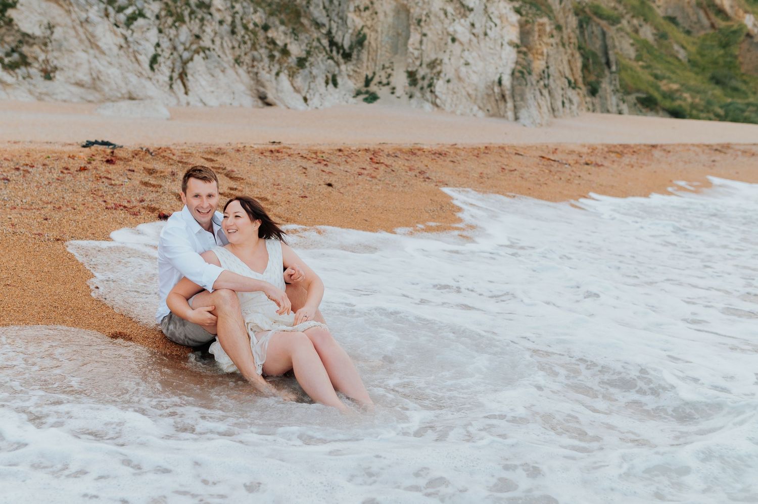 Two people share an intimate moment on a sandy beach near coastal rock formations.