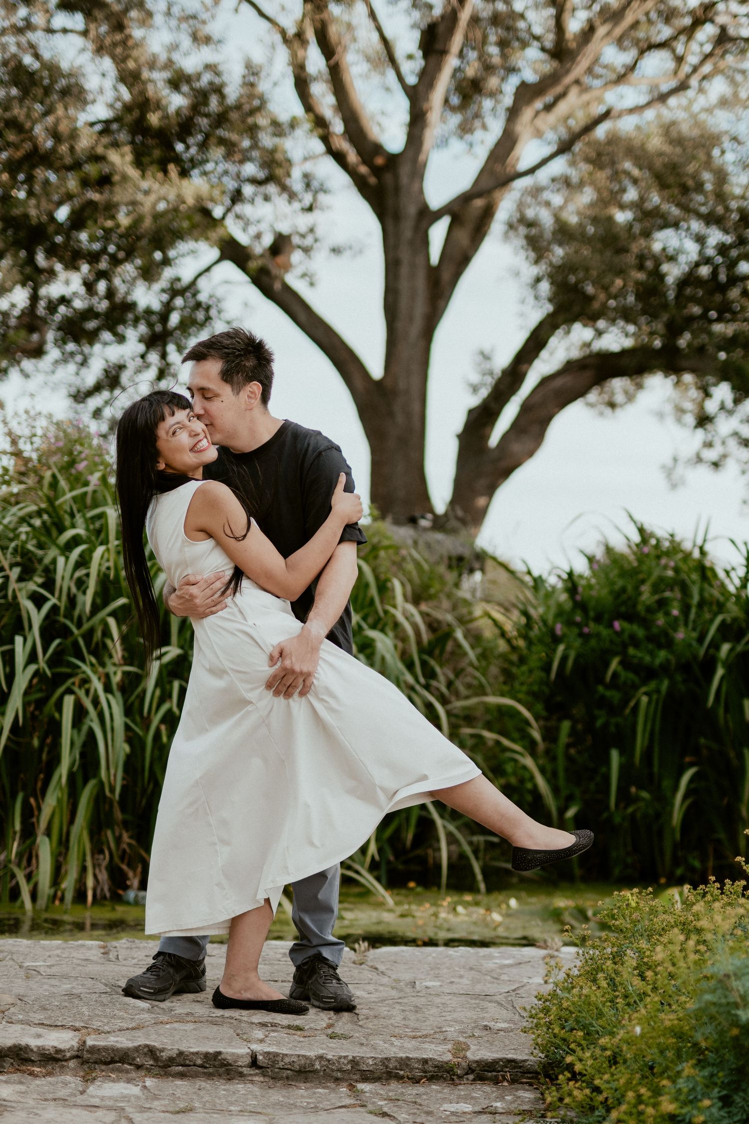 Playful couple poses in white dress and dark clothing under large trees in a garden.