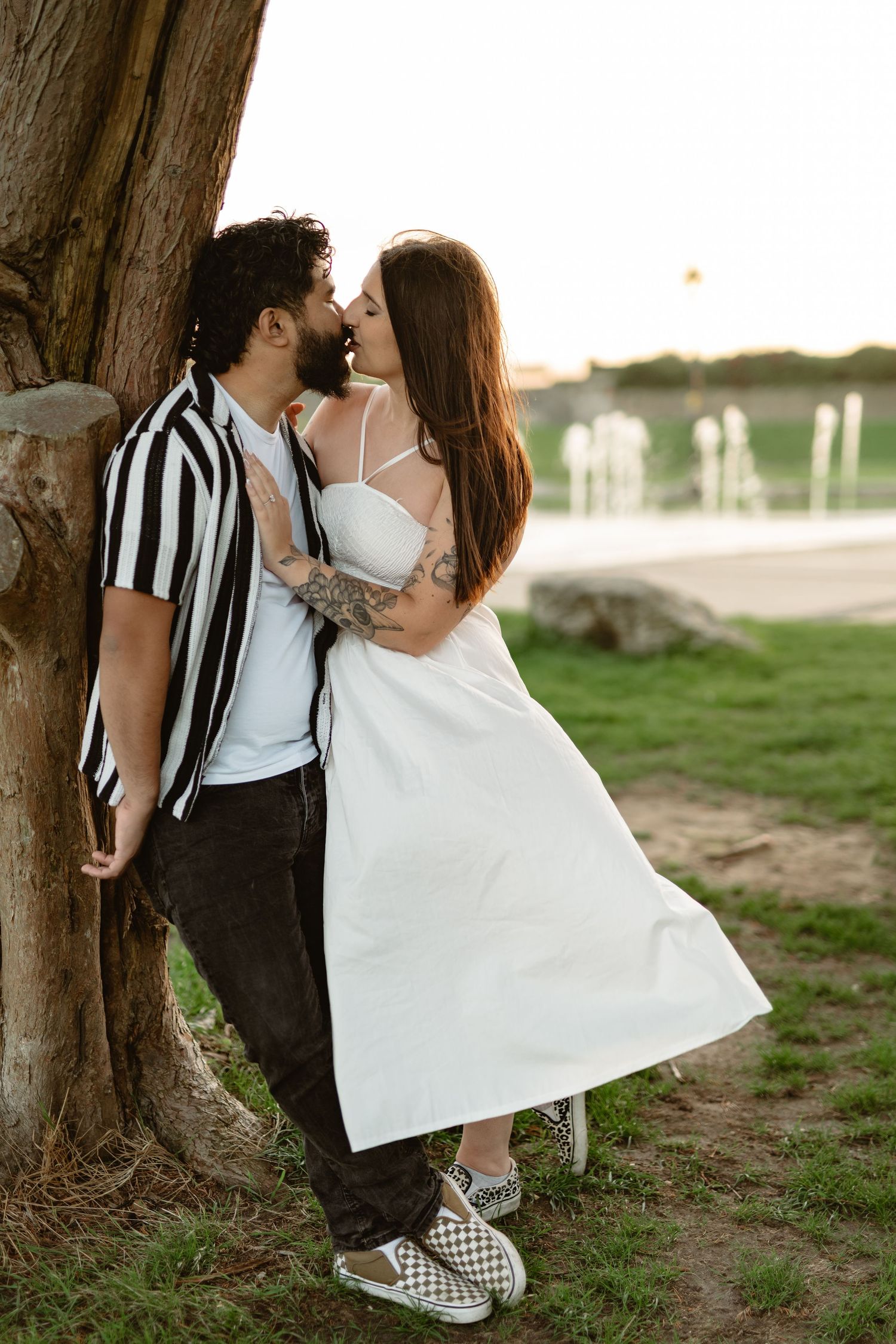 A couple in casual attire shares intimate moments near a large tree trunk.