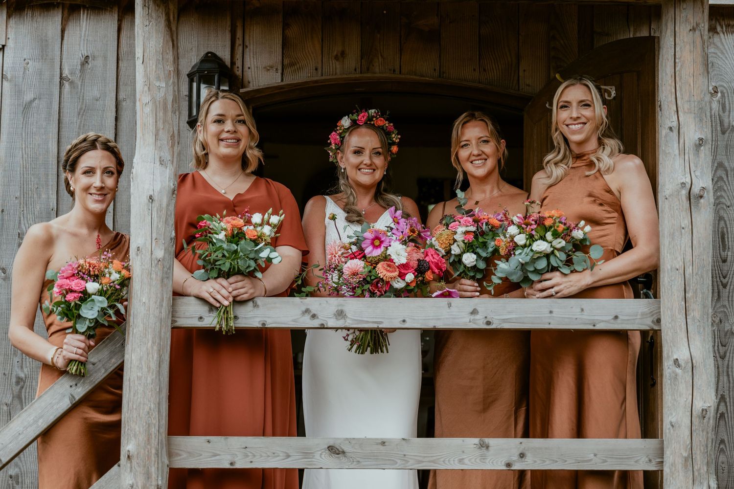 Bridesmaids in rust-colored dresses pose with pink bouquets on a rustic wooden porch.