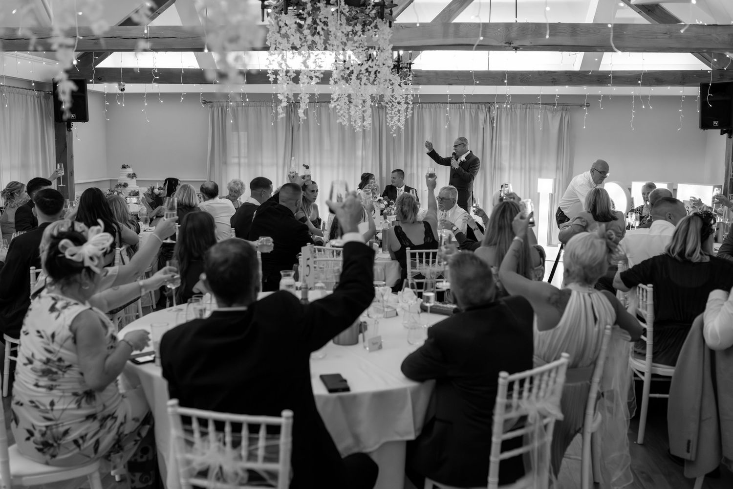 Black and white photo of wedding reception with guests seated at round tables under crystal chandeliers.