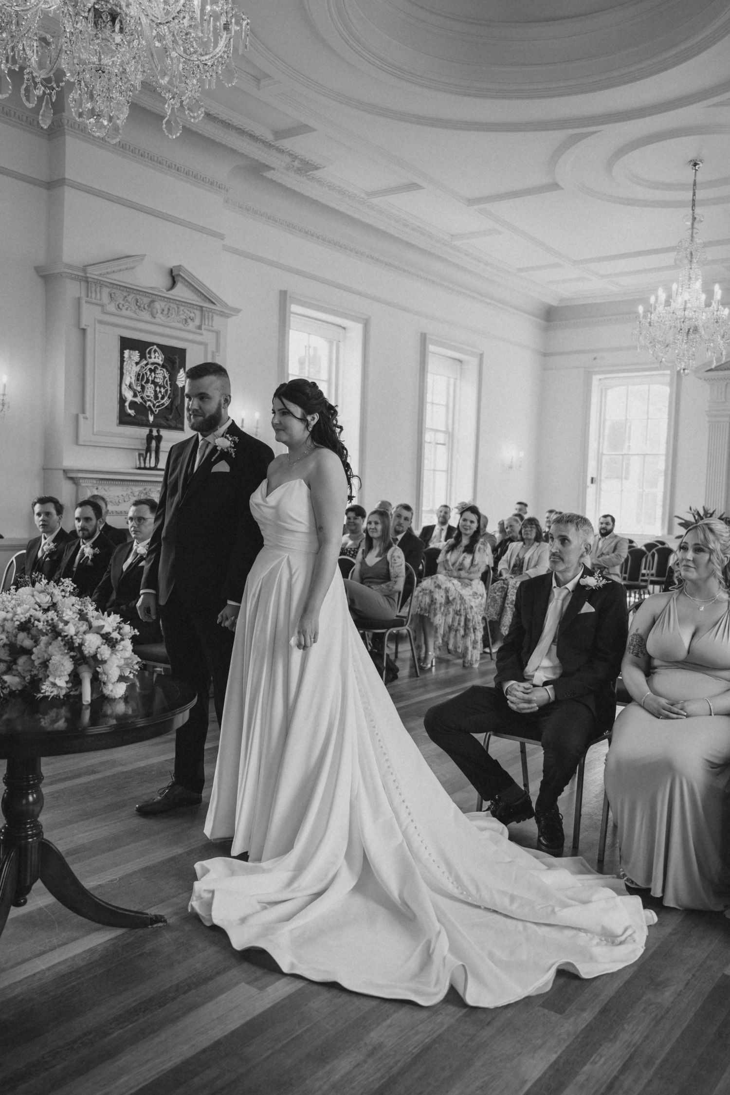 Wedding party dances in an ornate ballroom with crystal chandeliers and white decor.