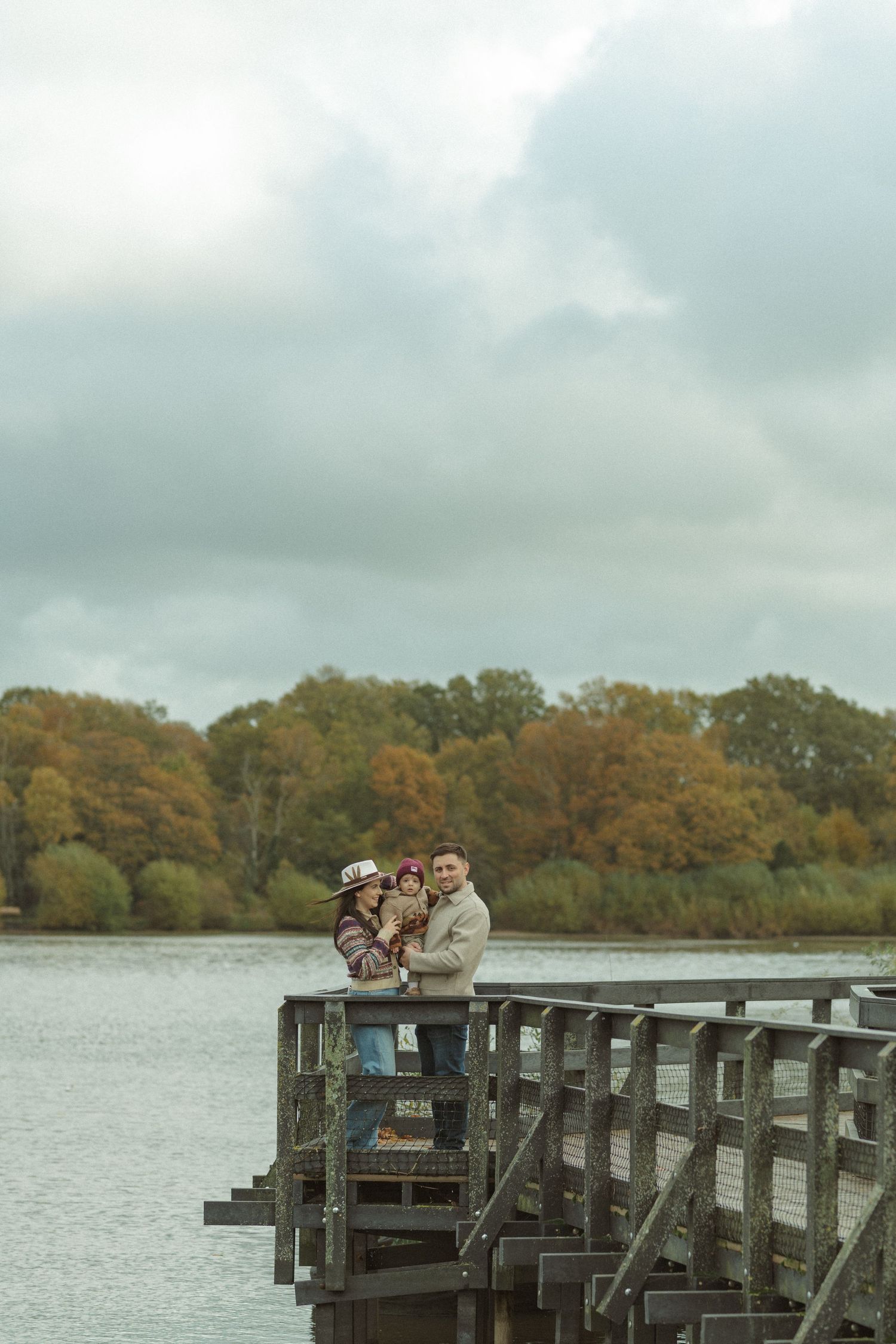 A family walks along a wooden pier over a lake surrounded by autumn trees with golden foliage.
