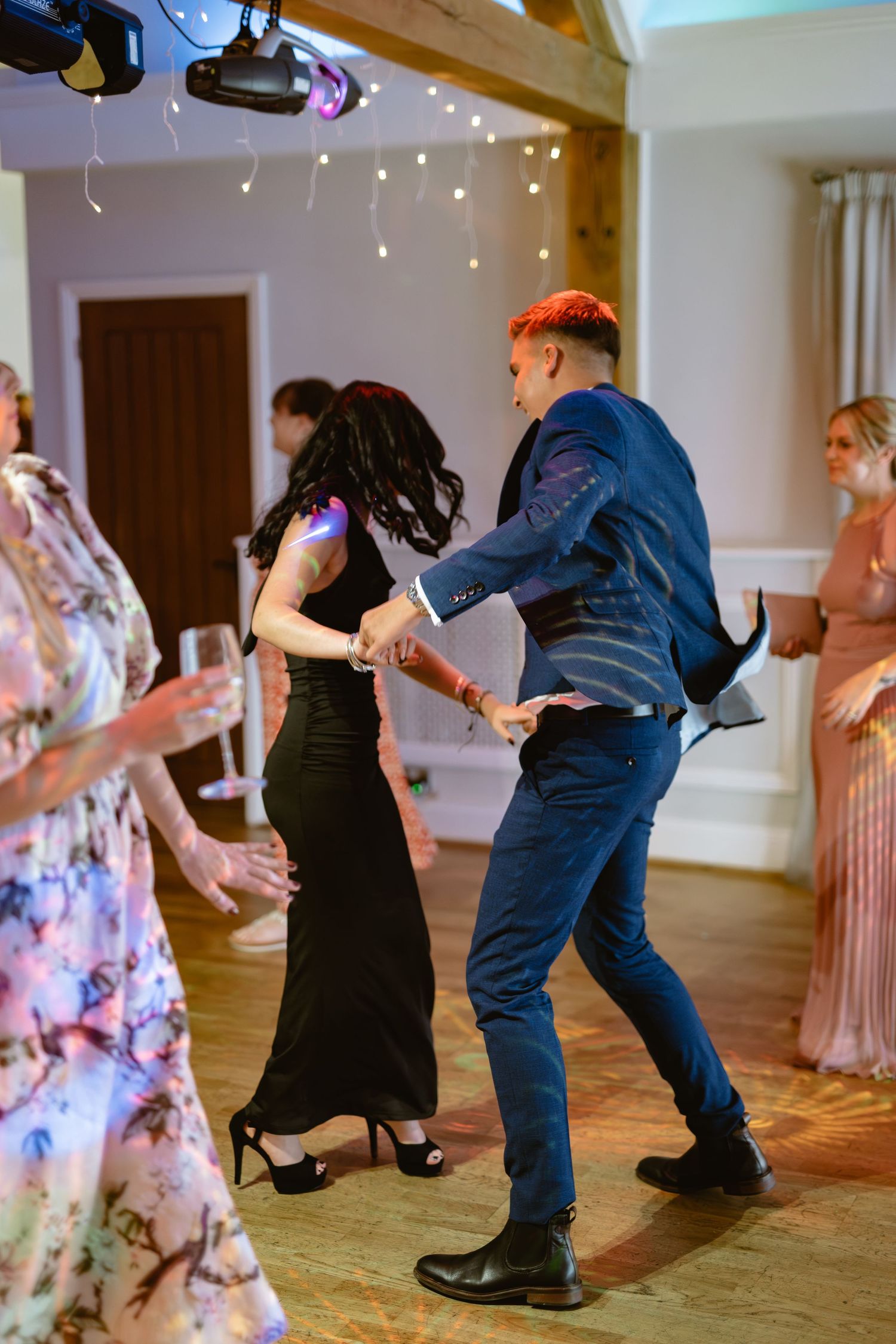 Wedding guests dancing enthusiastically on wooden dance floor under string lights.