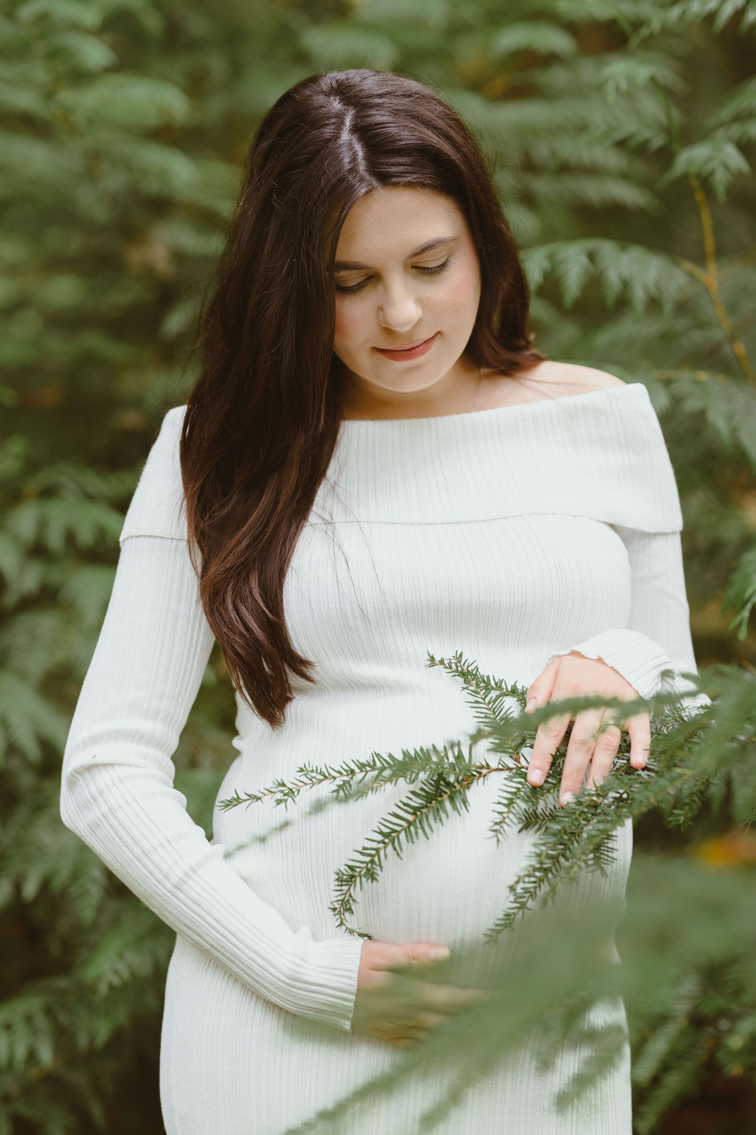 A person in white touches pine branches during a forest maternity photoshoot.