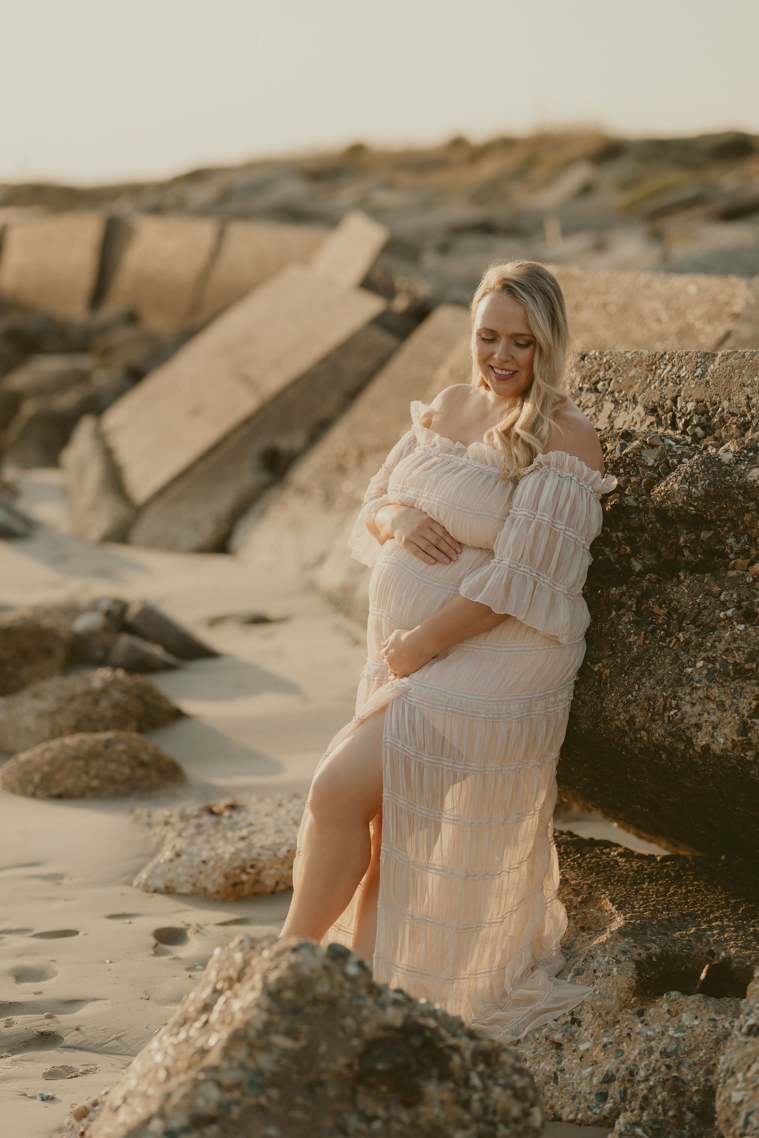 Maternity photo session on a rocky beach at sunset in a flowing white dress.