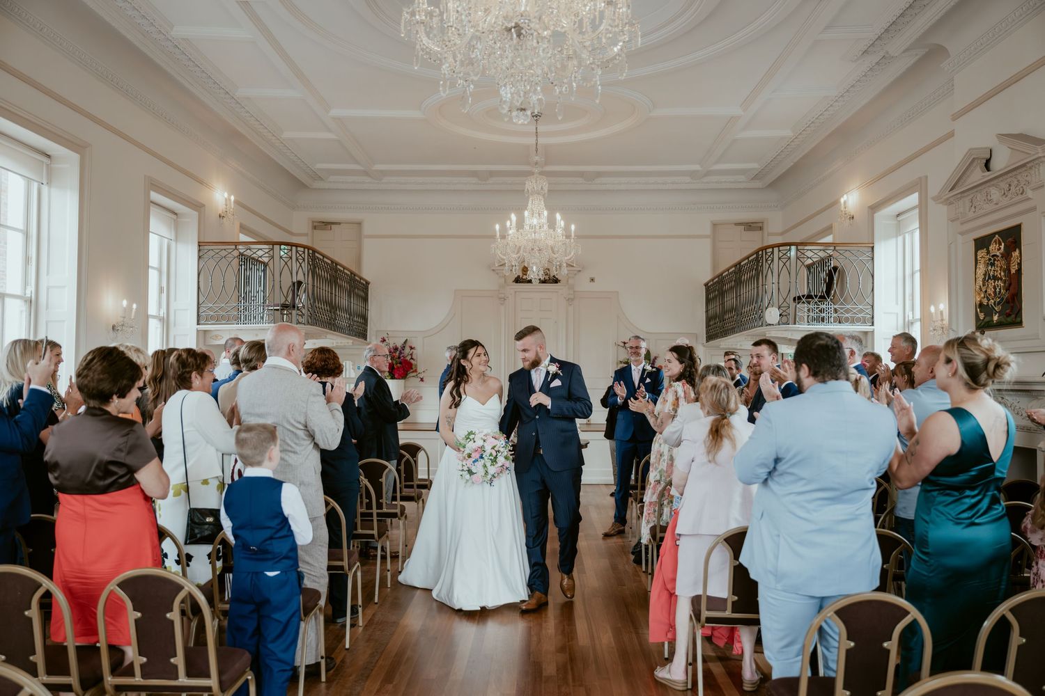 Guests celebrate during a wedding reception in a luxurious white ballroom with chandeliers.