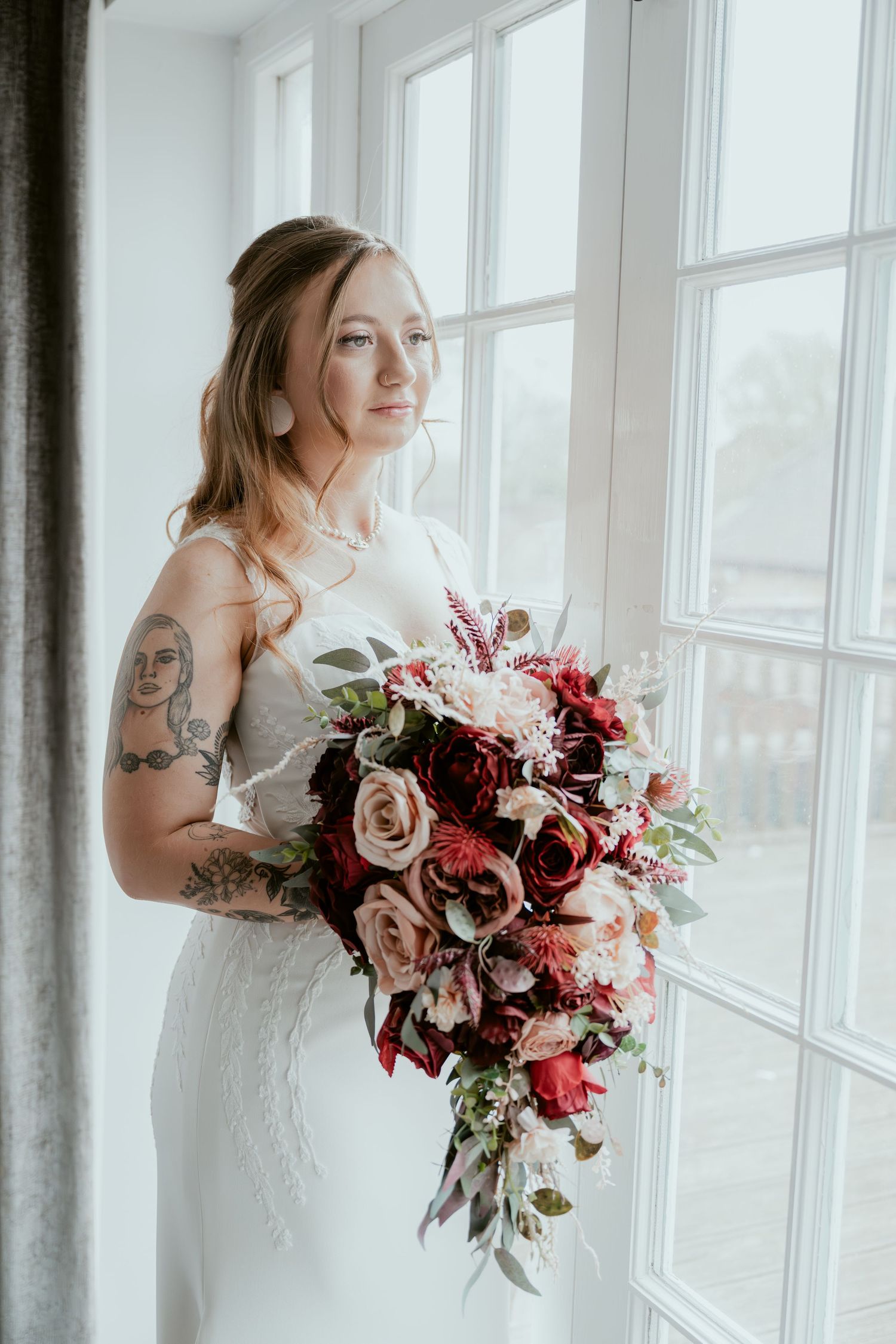 A bride in a white lace gown holds a stunning burgundy and blush rose bouquet by large windows.