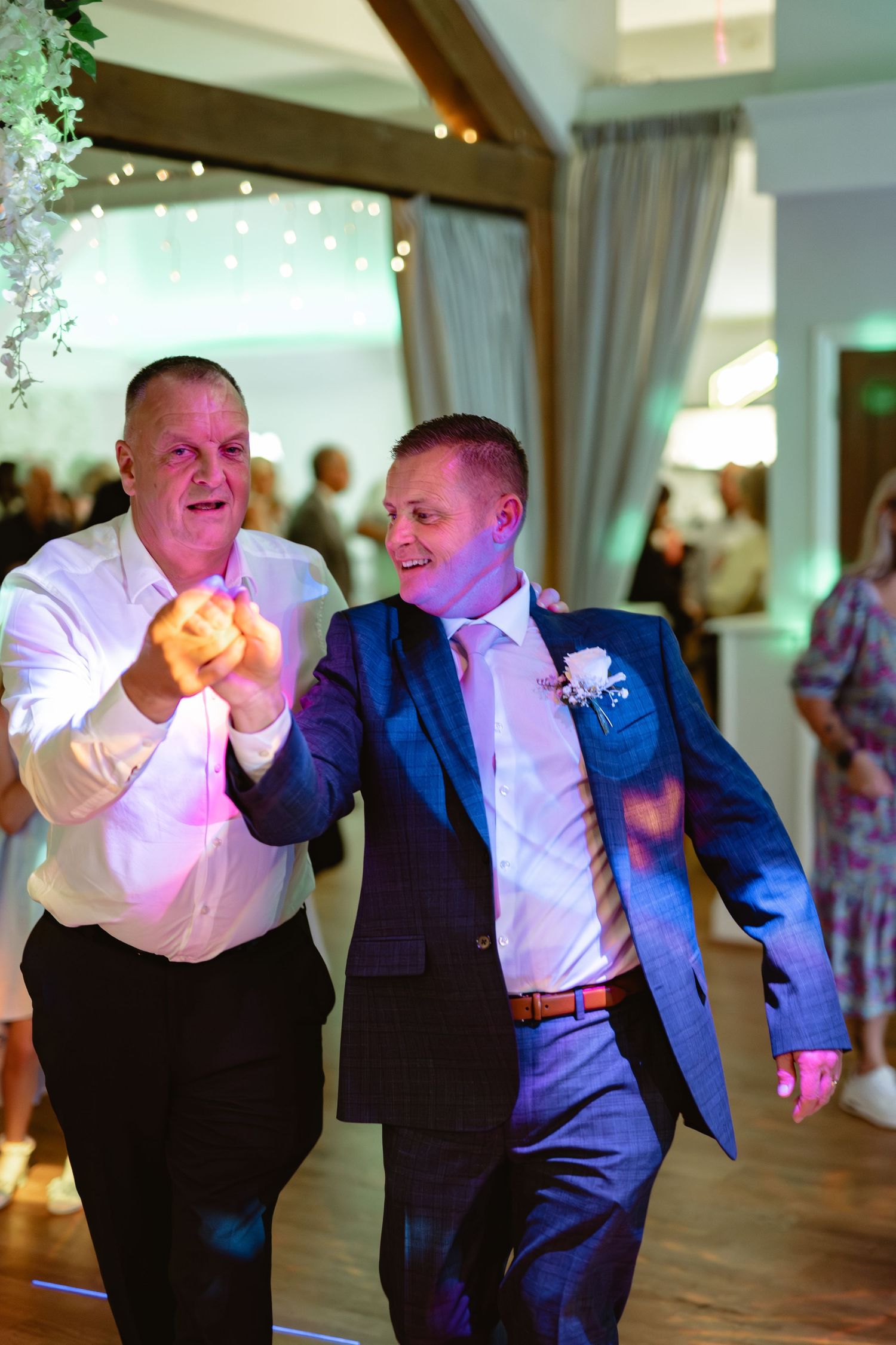 Wedding guests perform energetic dance moves in formal attire at indoor reception.