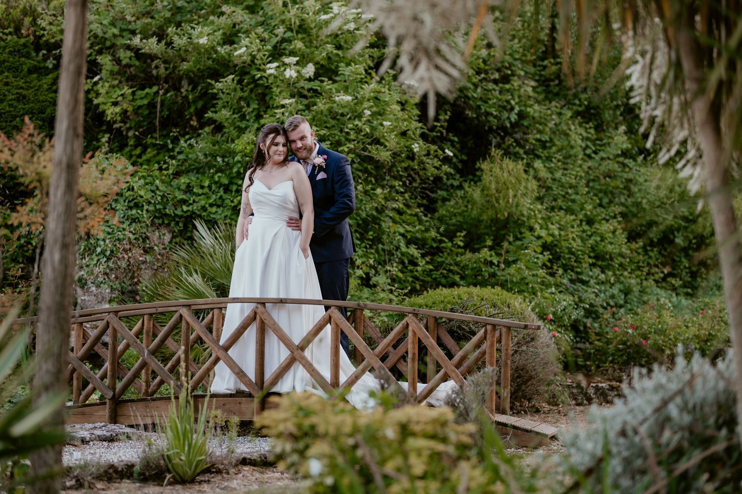 Wedding couple walking along rustic wooden bridge surrounded by lush gardens.