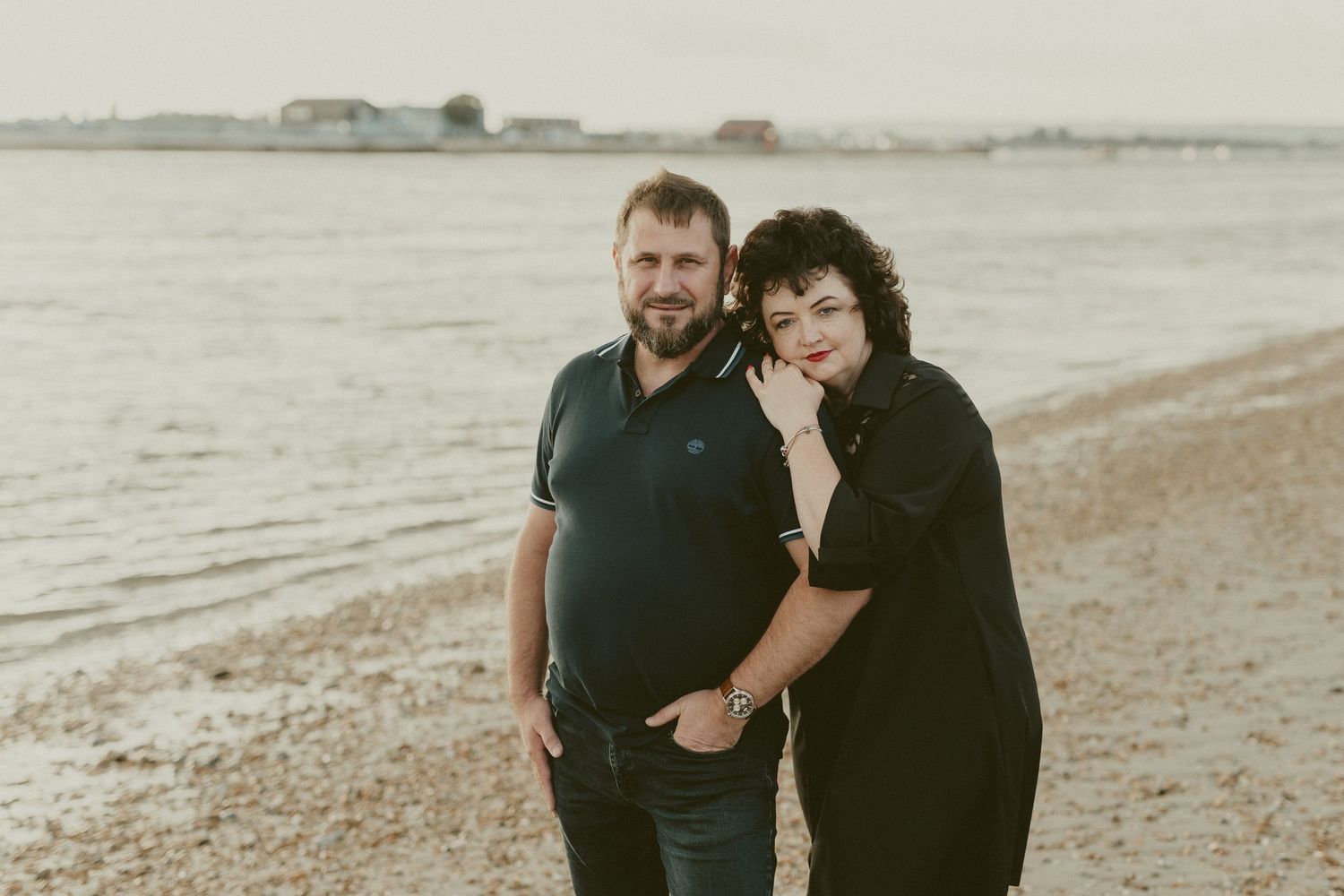 A couple poses casually on a pebble beach during golden hour.