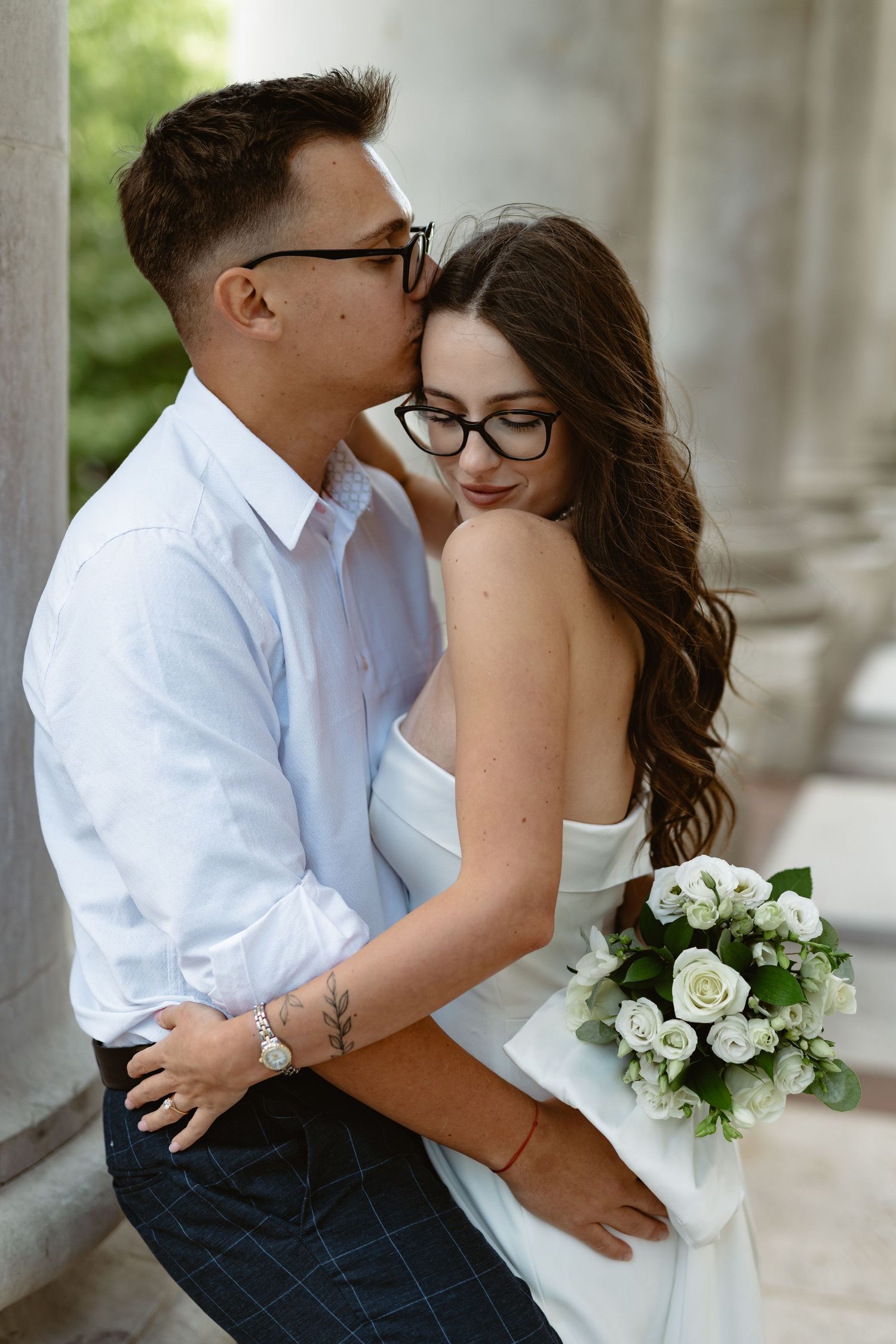Couple in coordinated outfits share tender moments near classical columns with white bouquet.