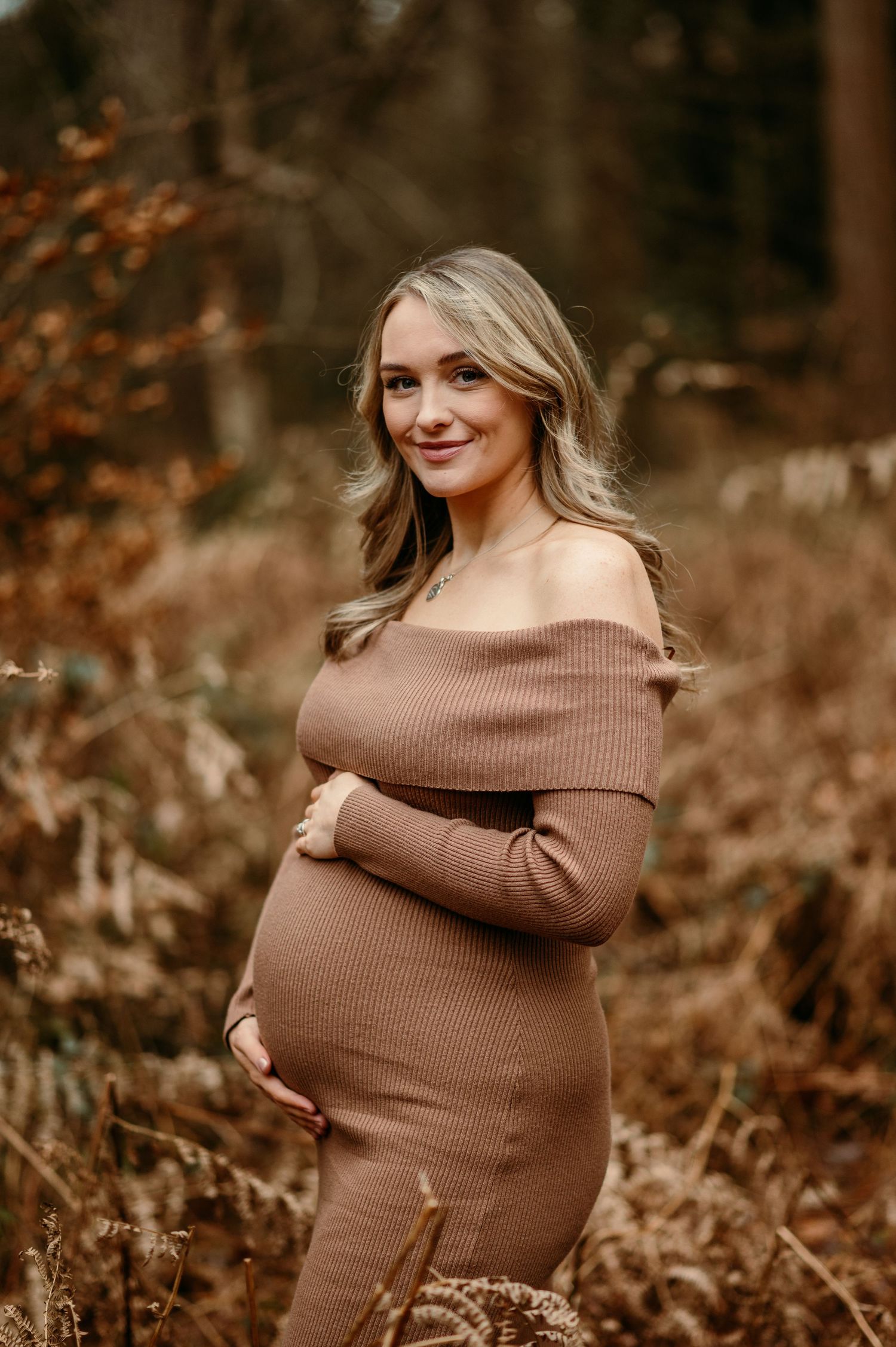 Expectant mother in a brown sweater dress poses joyfully in an autumn forest setting.