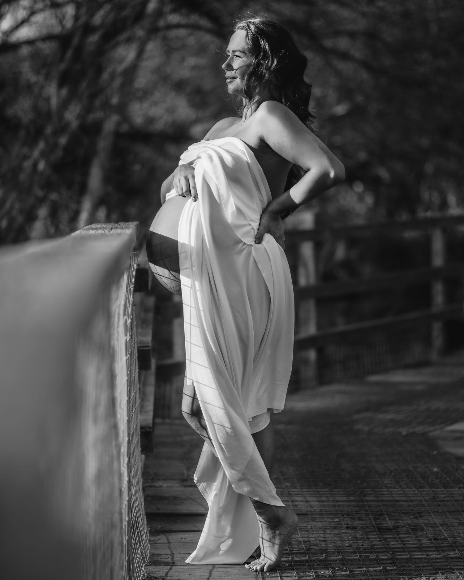 Artistic black and white maternity photos showing figure in white dress leaning against wooden railing.
