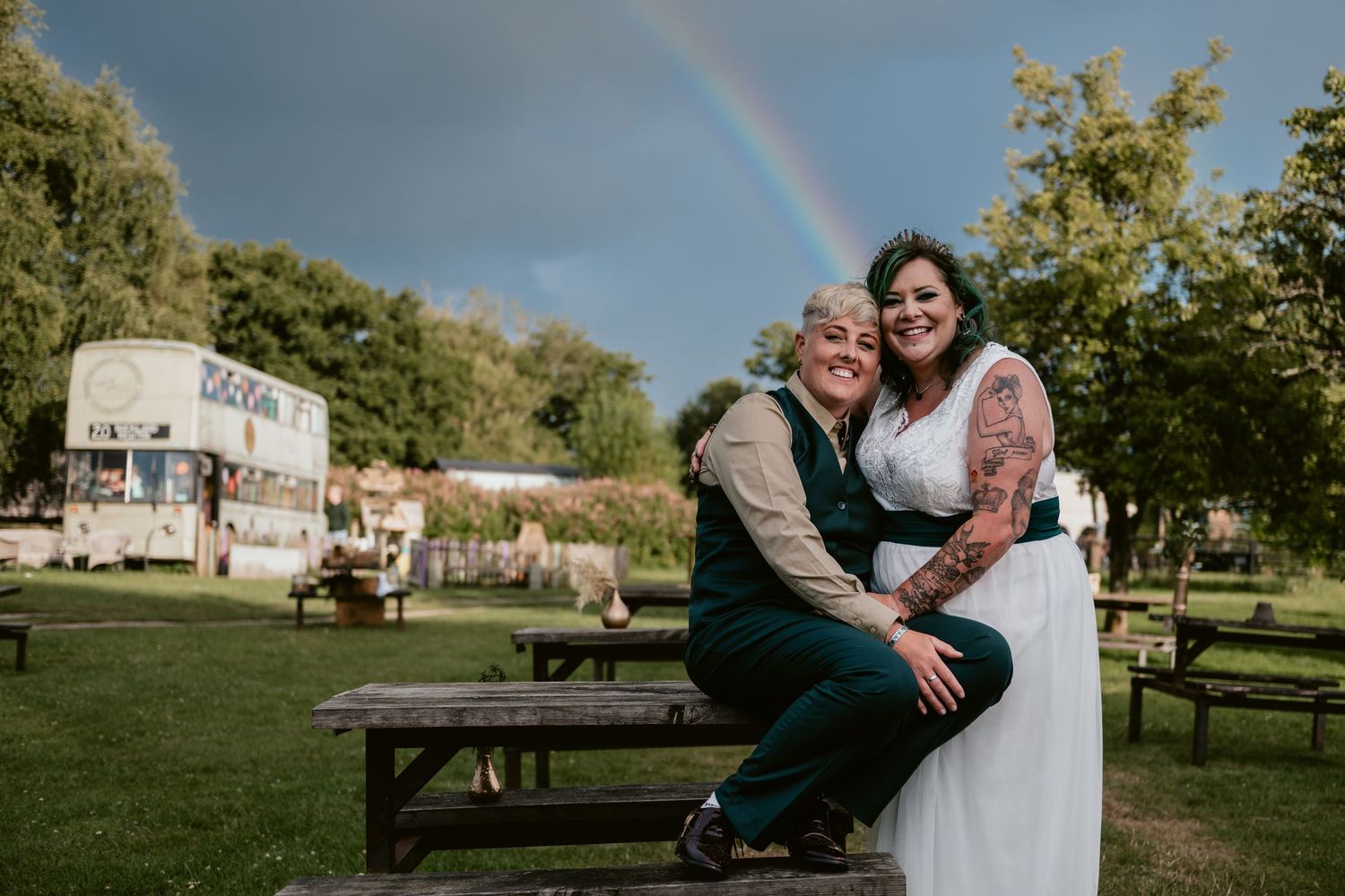 A couple sits together on a picnic bench with a rainbow and vintage camper van in the background.