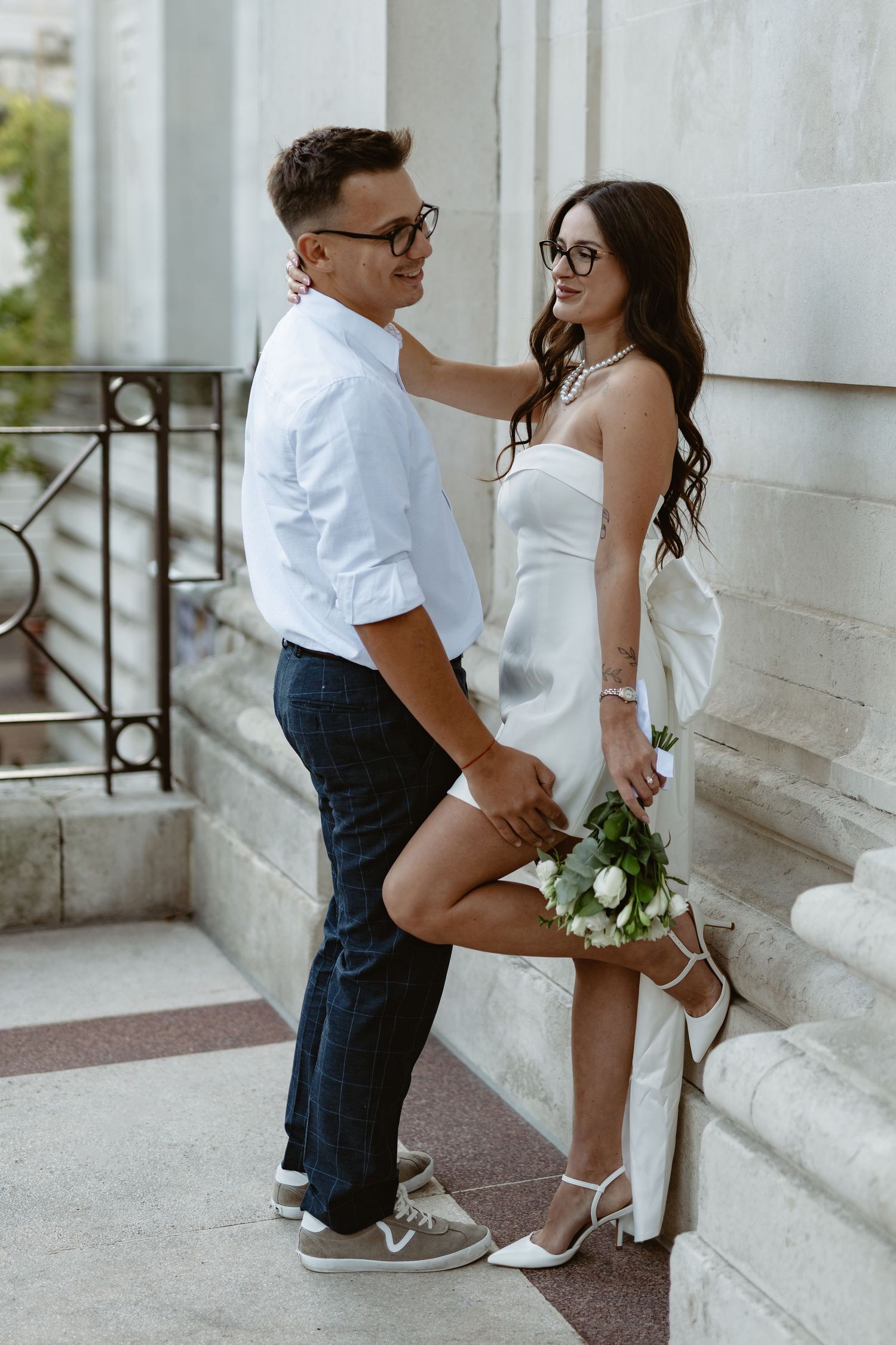 Couple in white outfits posing playfully against white building wall.