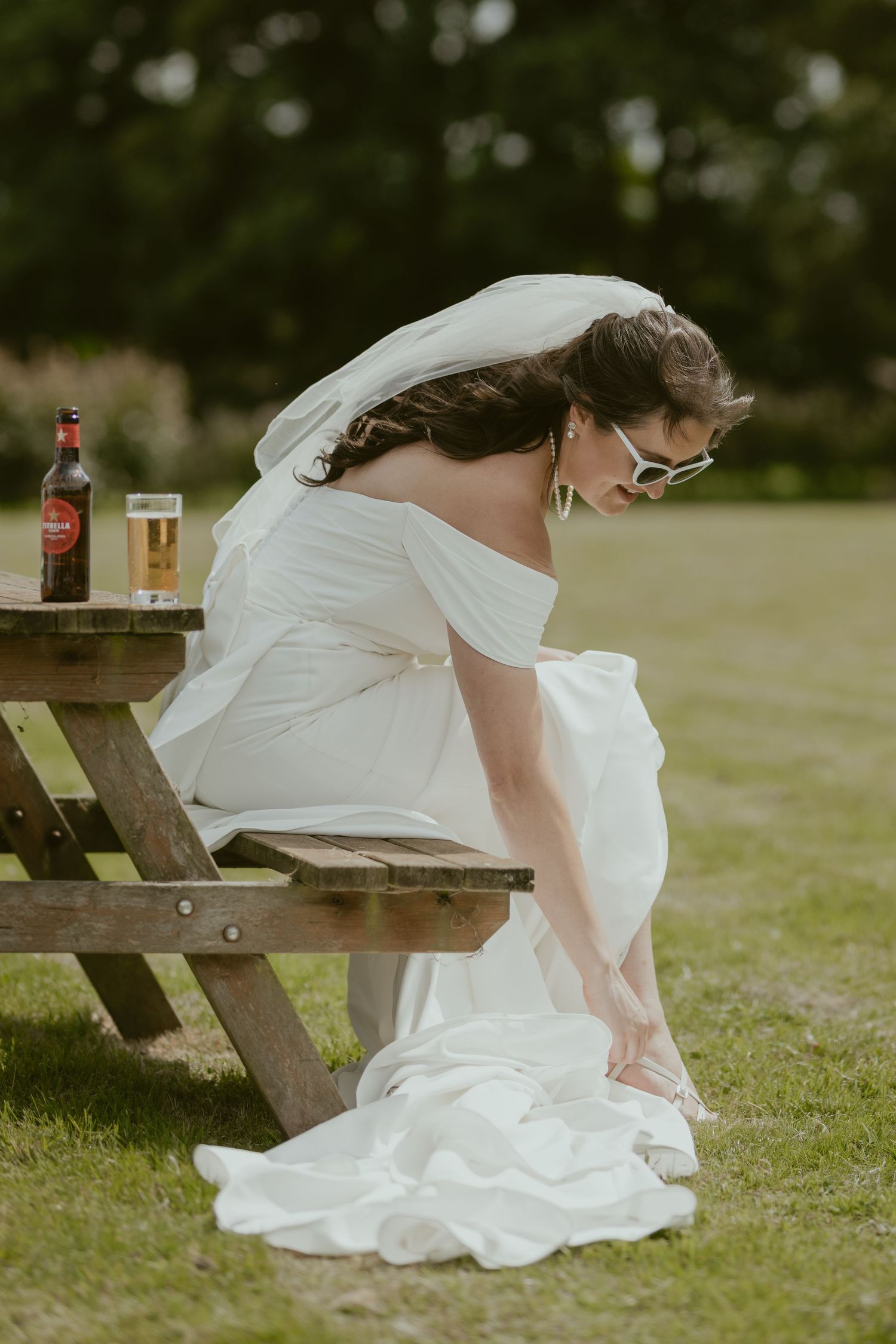 Bride in white dress sitting at picnic table with refreshments.