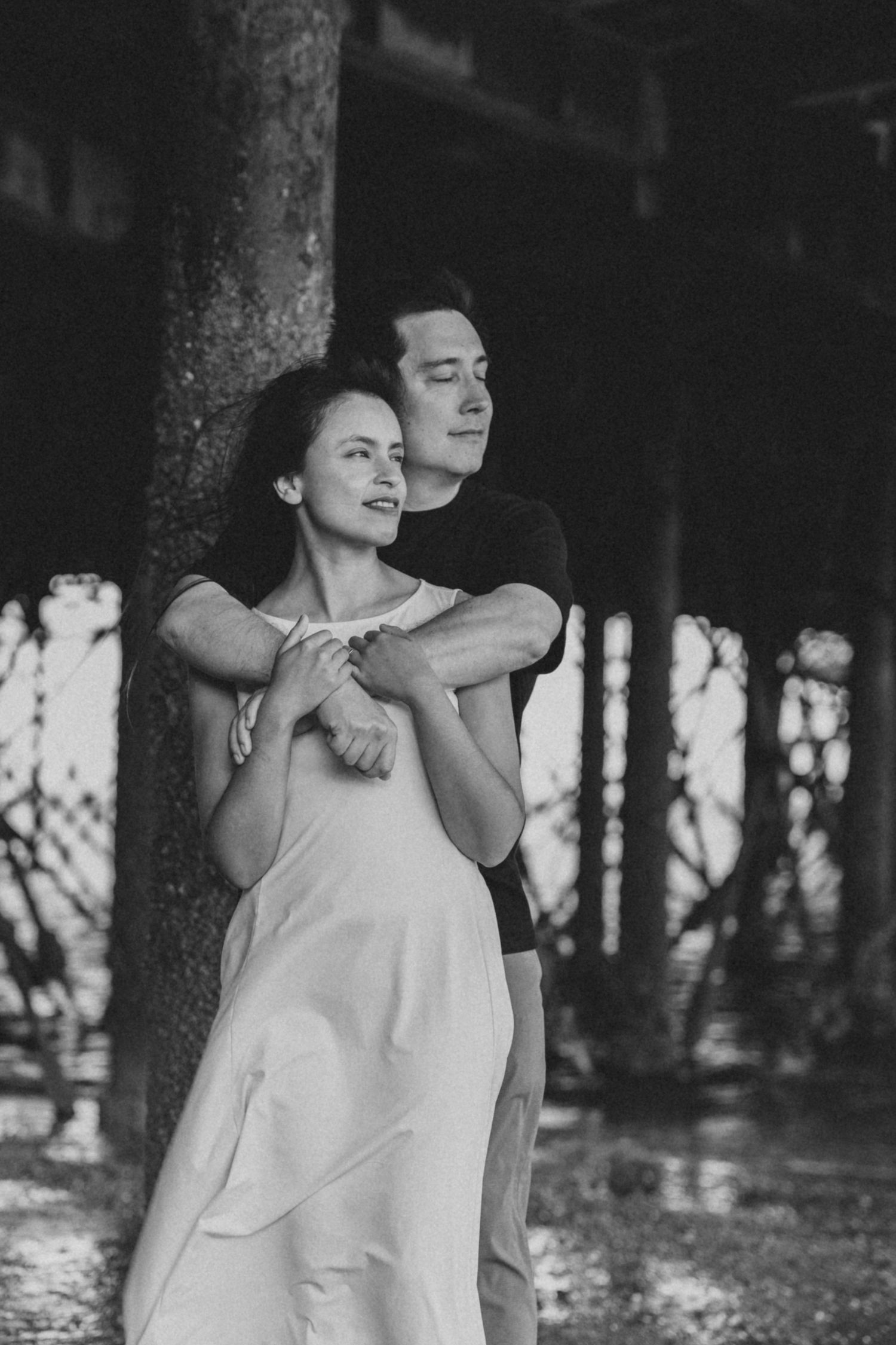 A romantic black and white photo of a couple embracing among trees in a forest setting.