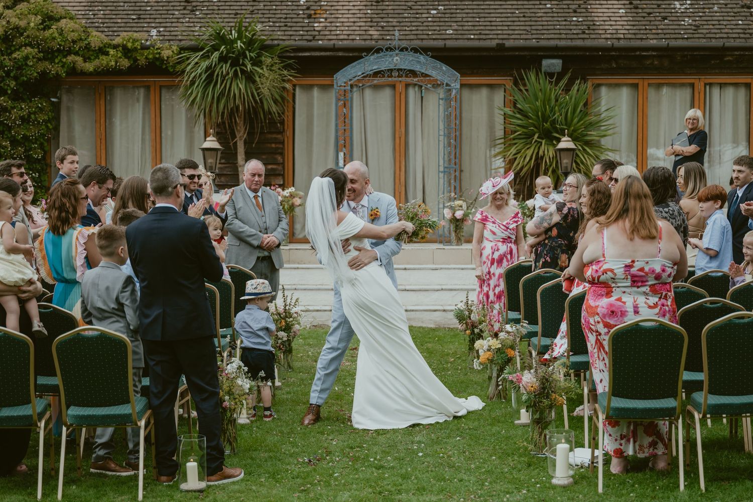 Wedding guests seated in white chairs on a lawn in front of a stone building.