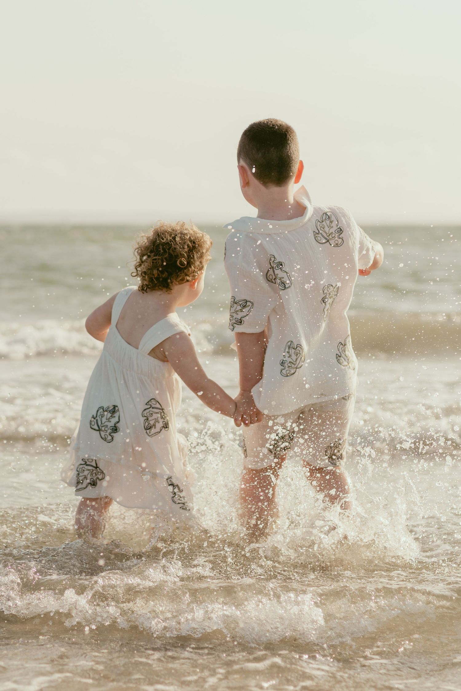 Two people in white outfits splash and play in ocean waves.