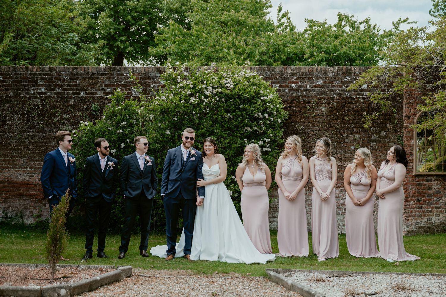 Wedding party poses for photos in a garden setting with brick walls and greenery.
