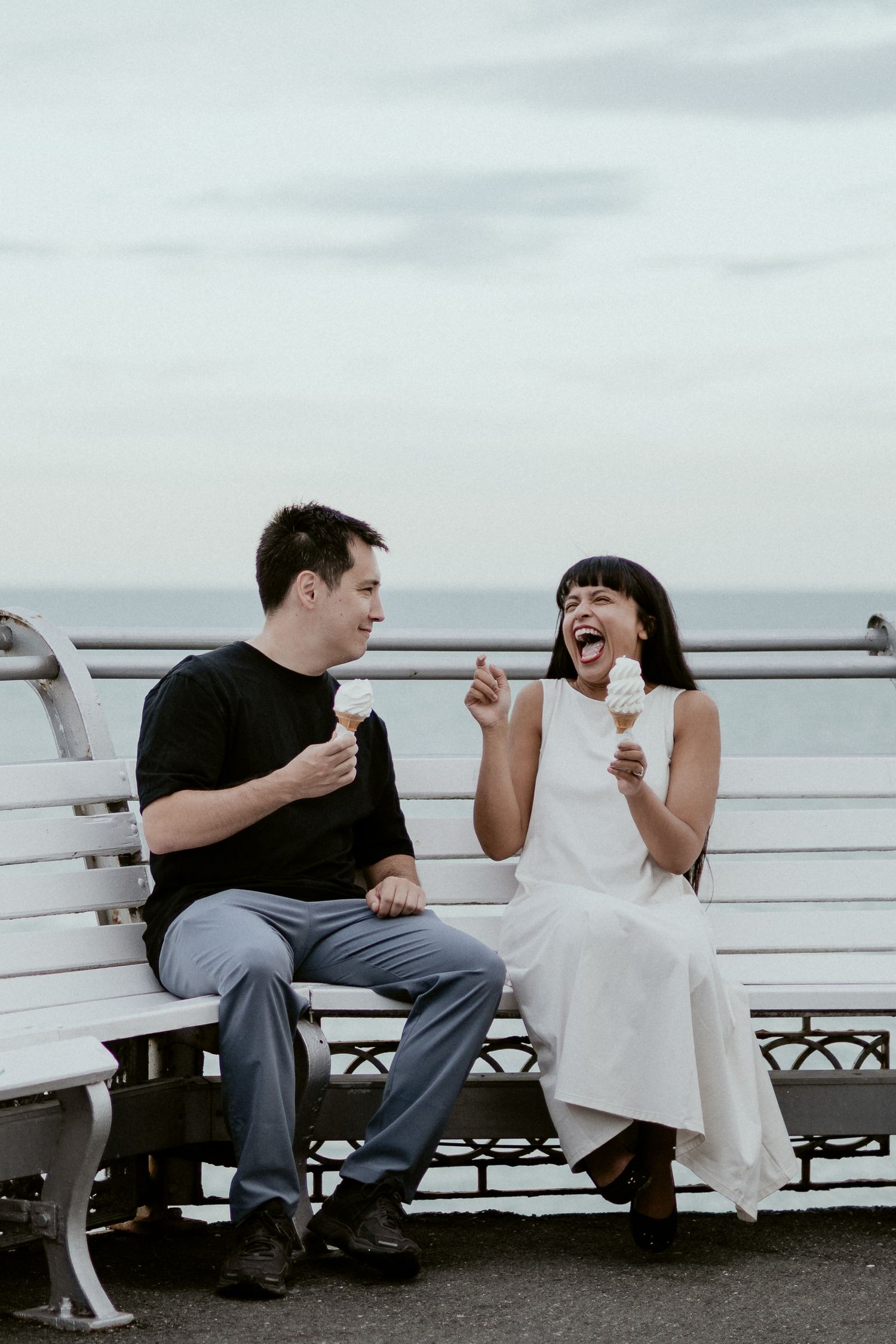 Two people share ice cream cones while sitting on a seaside bench and laughing together.