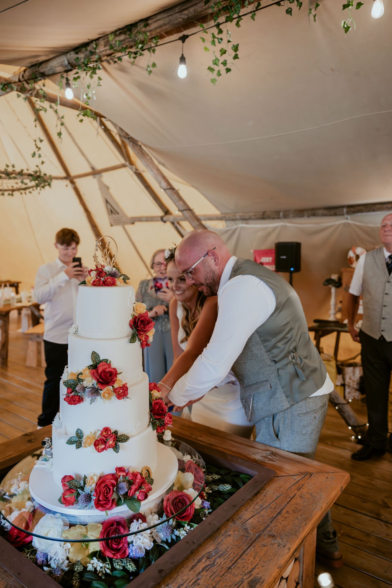 Wedding cake with colorful floral decorations being cut on rustic wooden table inside tent.