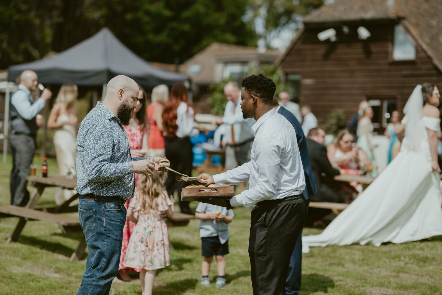 Outdoor wedding reception with guests gathered near a rustic wooden building.