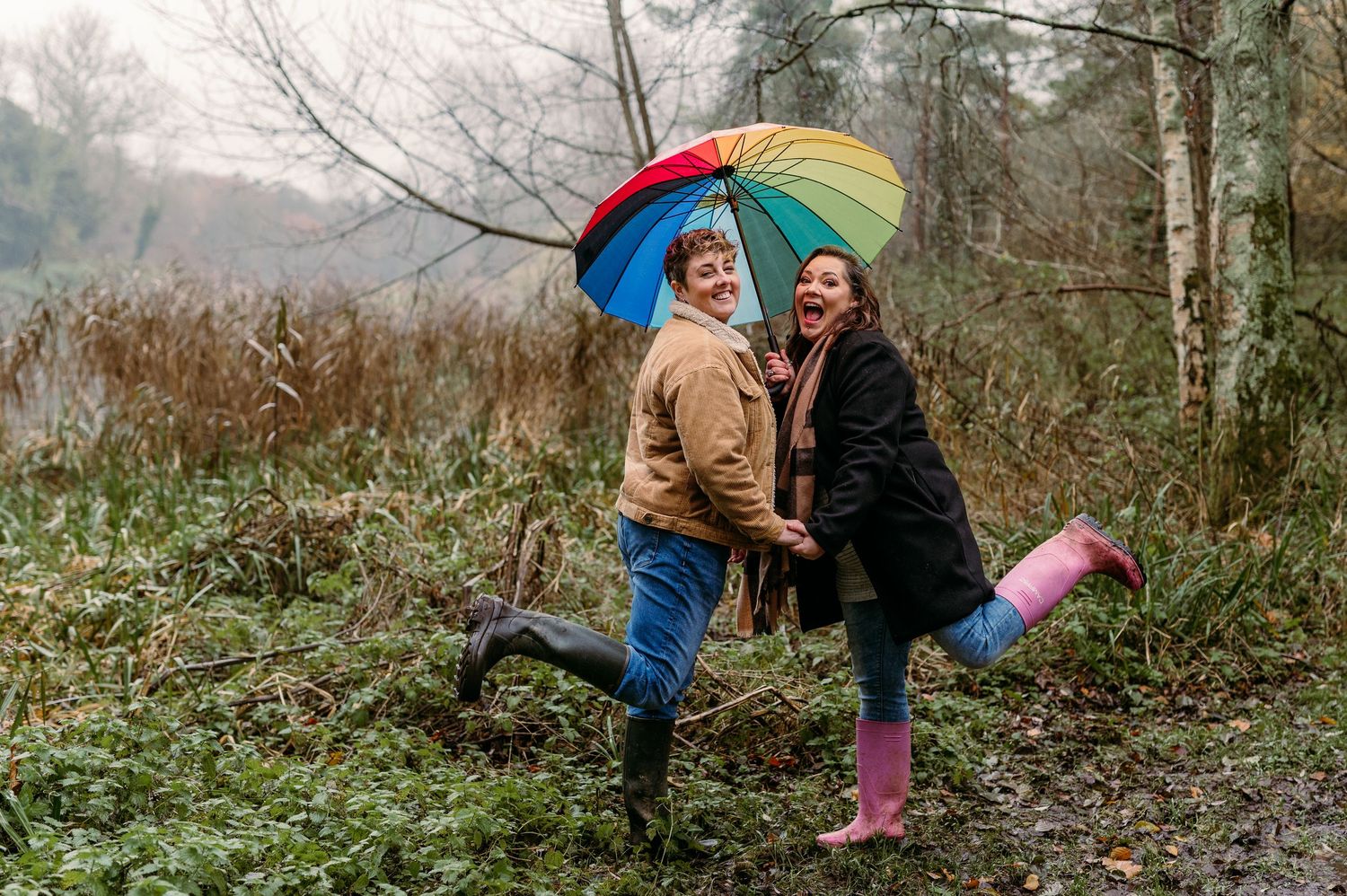 Couple walking through autumn woods with a colorful rainbow umbrella.