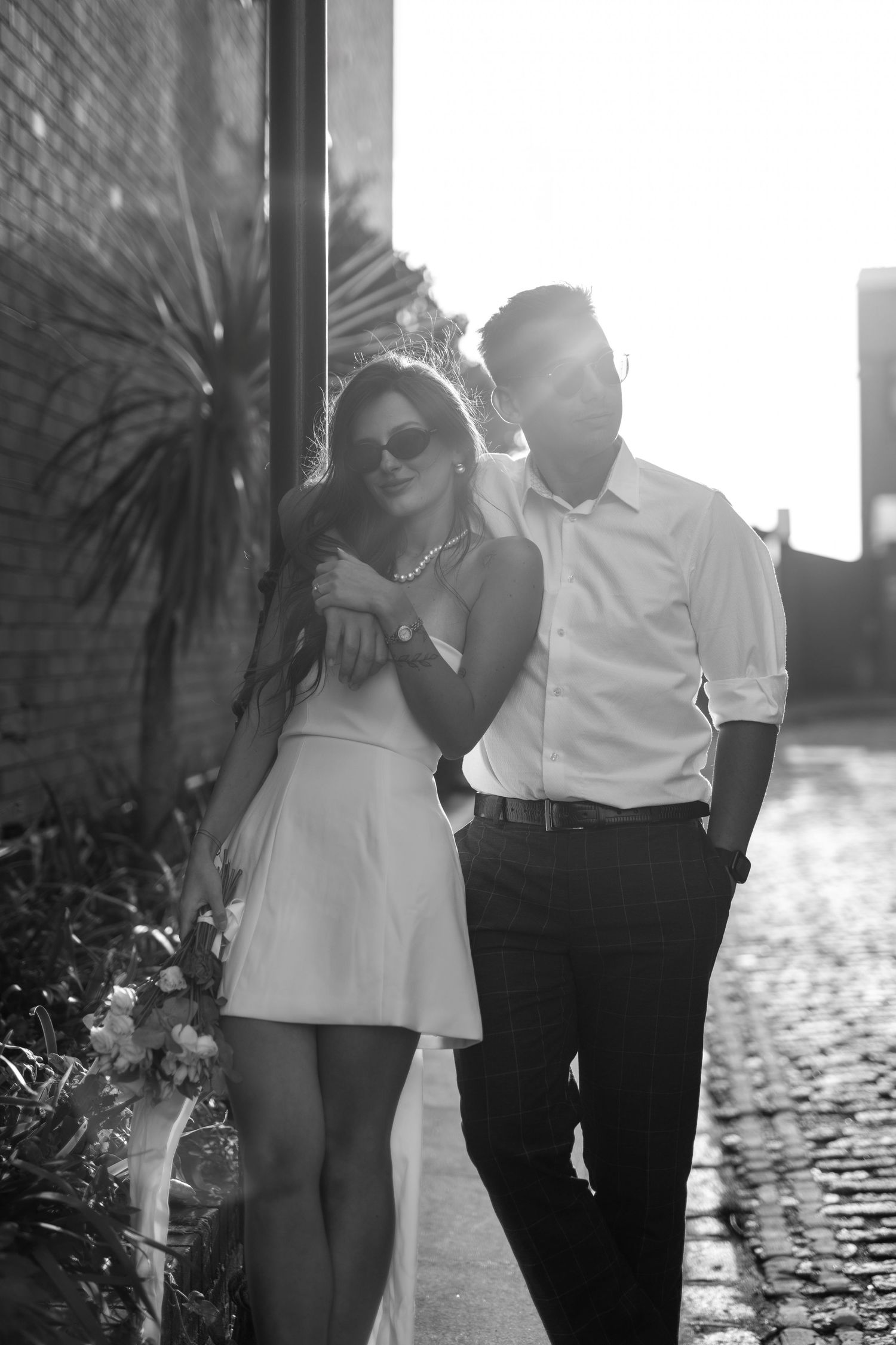 Sun-lit black and white photo series of a couple in white and dark clothing against a brick wall.