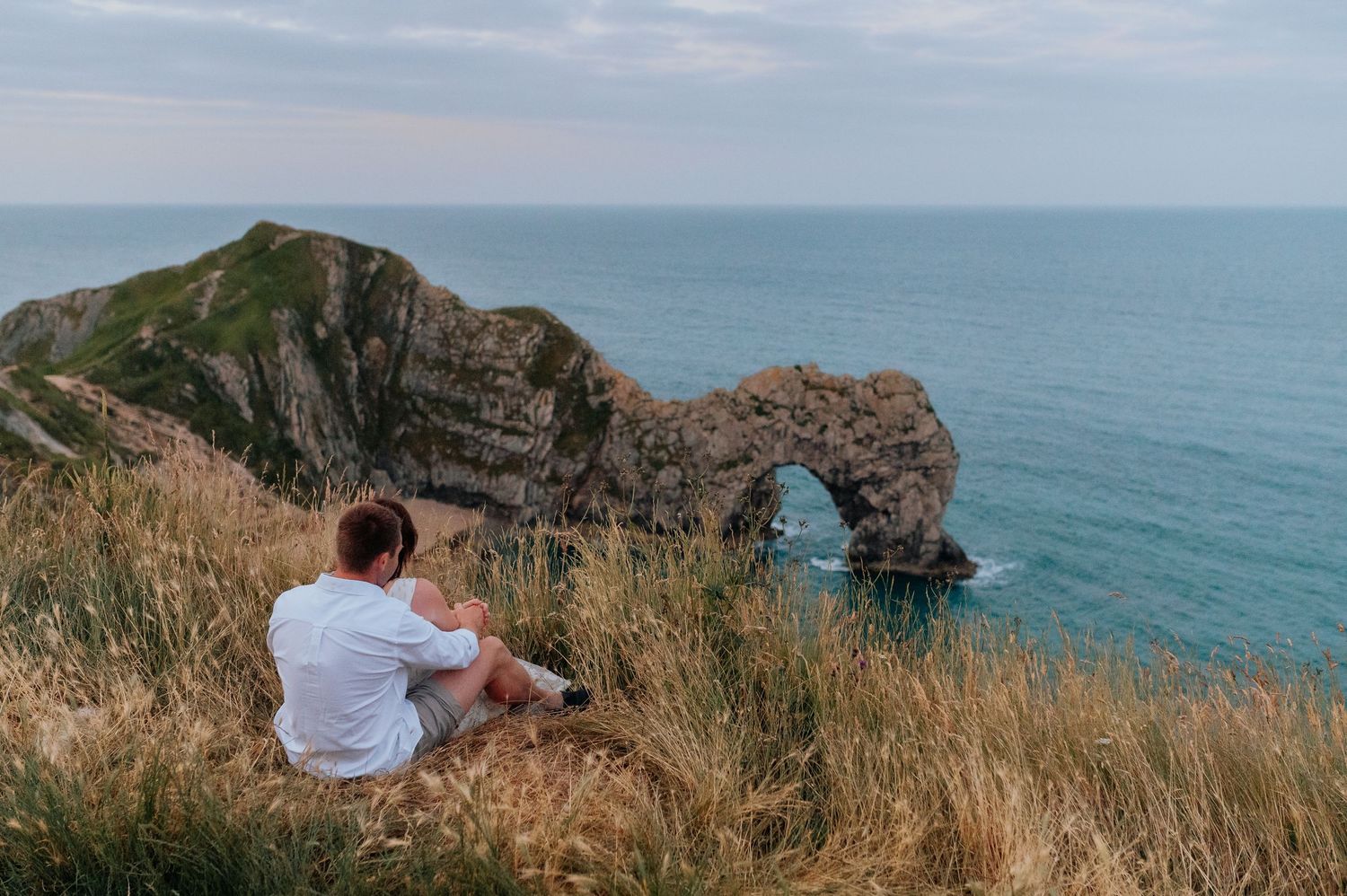Two people sit on coastal cliffs overlooking a beautiful ocean landscape.