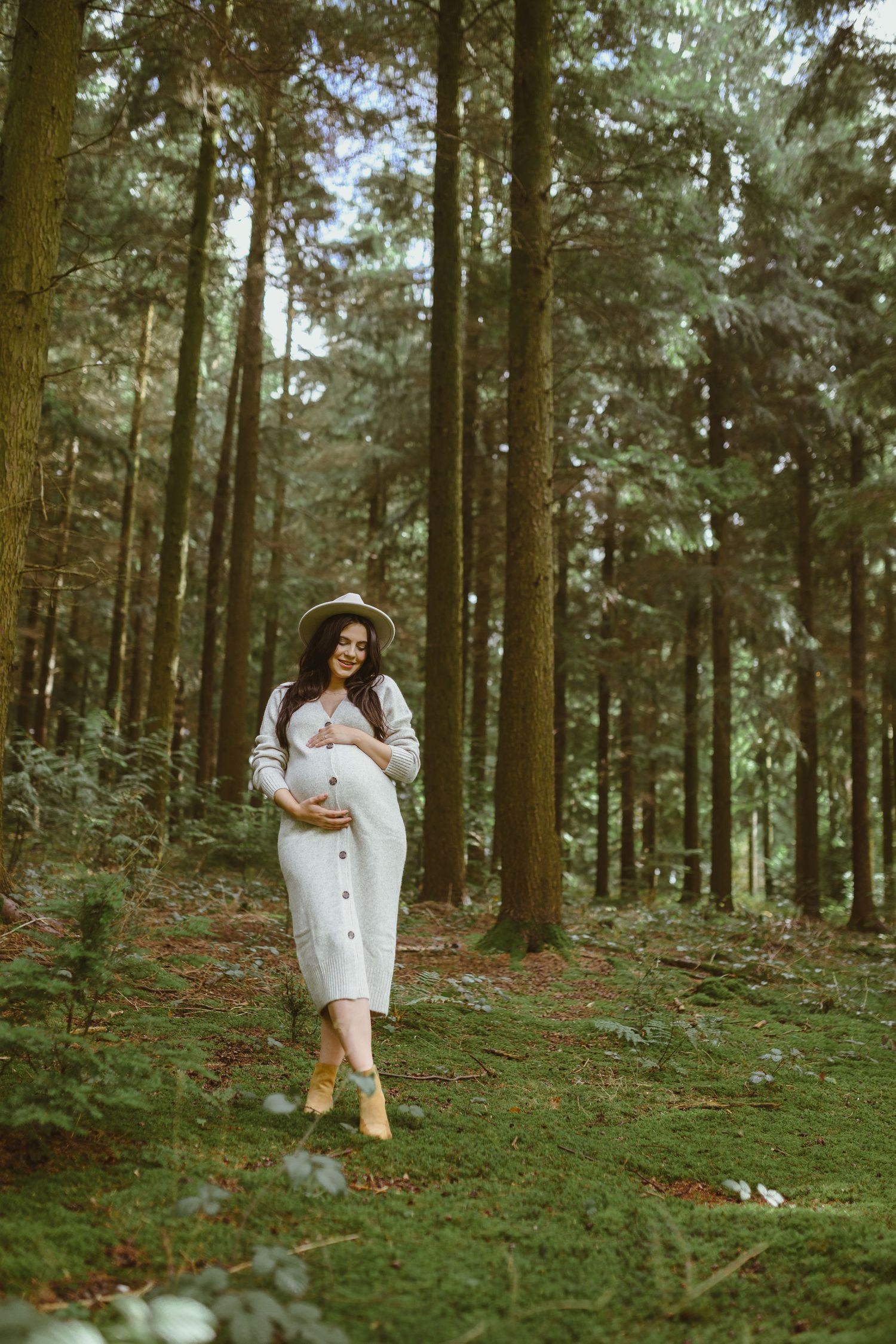 A maternity photoshoot in a lush forest setting with a person wearing a white sweater dress and brown hat.