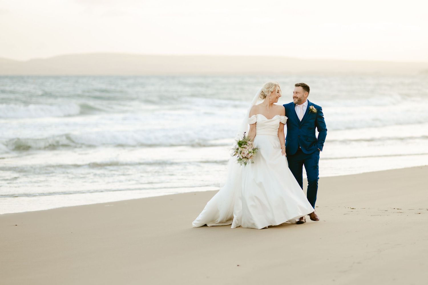 Couple takes romantic photos along sandy beach at sunset.