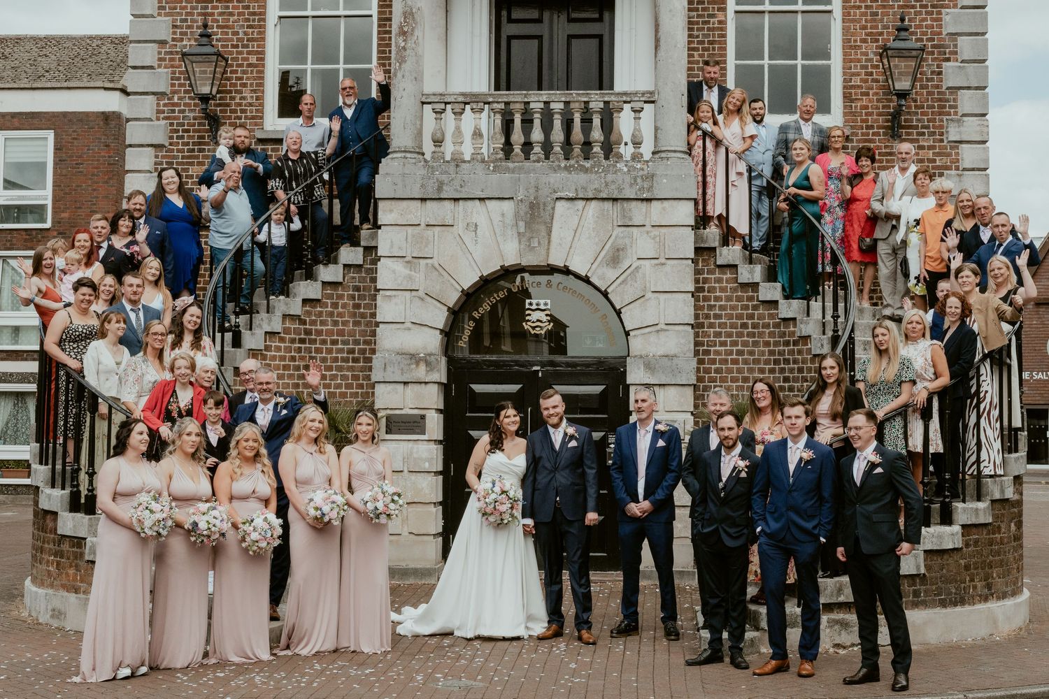 Wedding party poses for formal photos on the steps of a historic brick building.