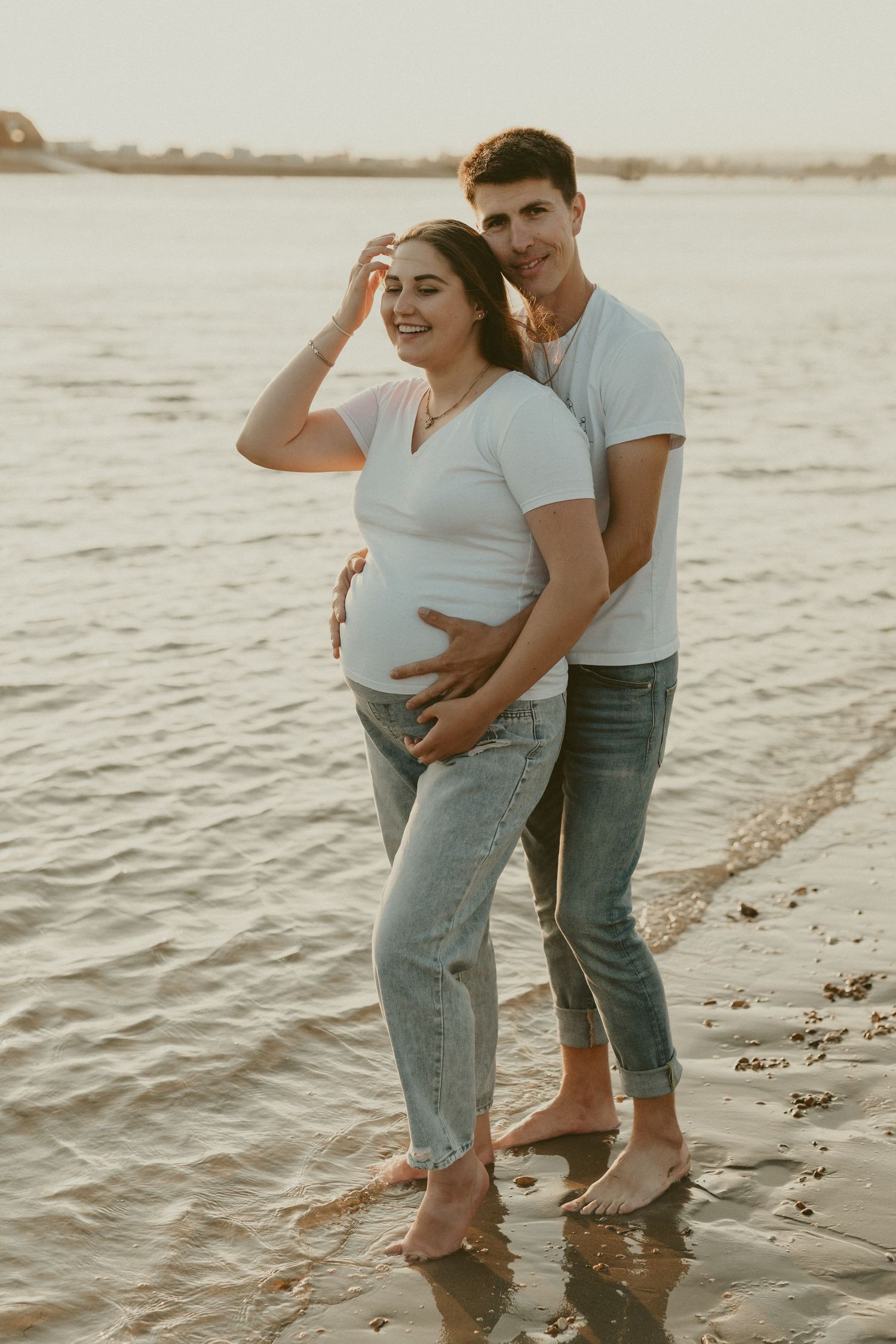 Couple standing in shallow water at sunset wearing casual denim and white shirts.