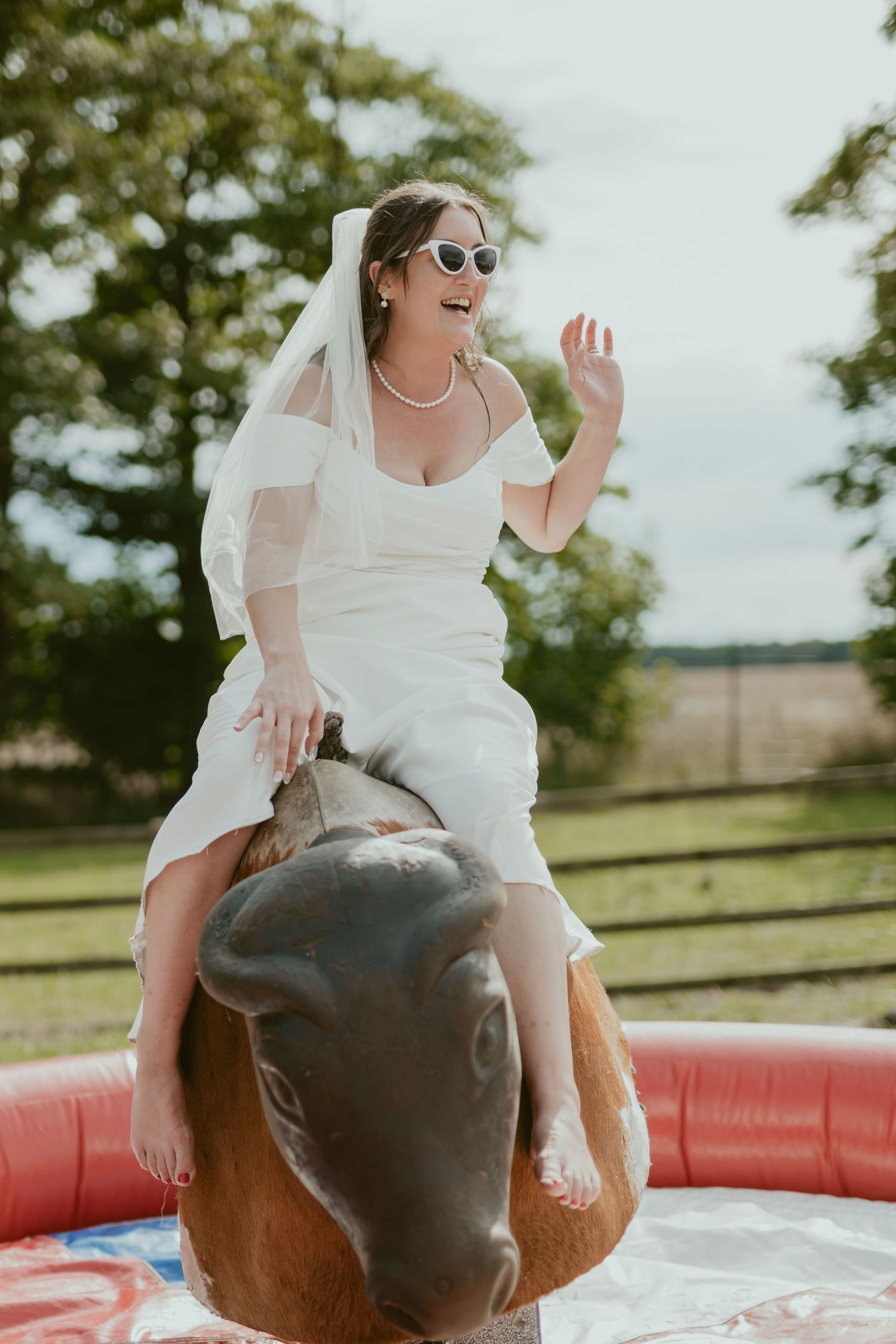 Bride rides mechanical bull in wedding dress while wearing sunglasses.