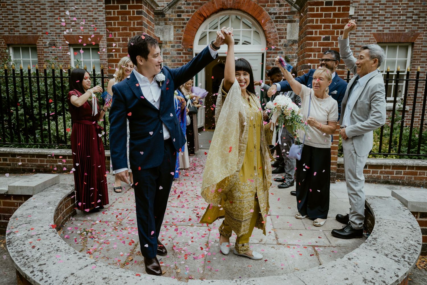 Wedding celebration outside a brick church with guests throwing pink confetti.