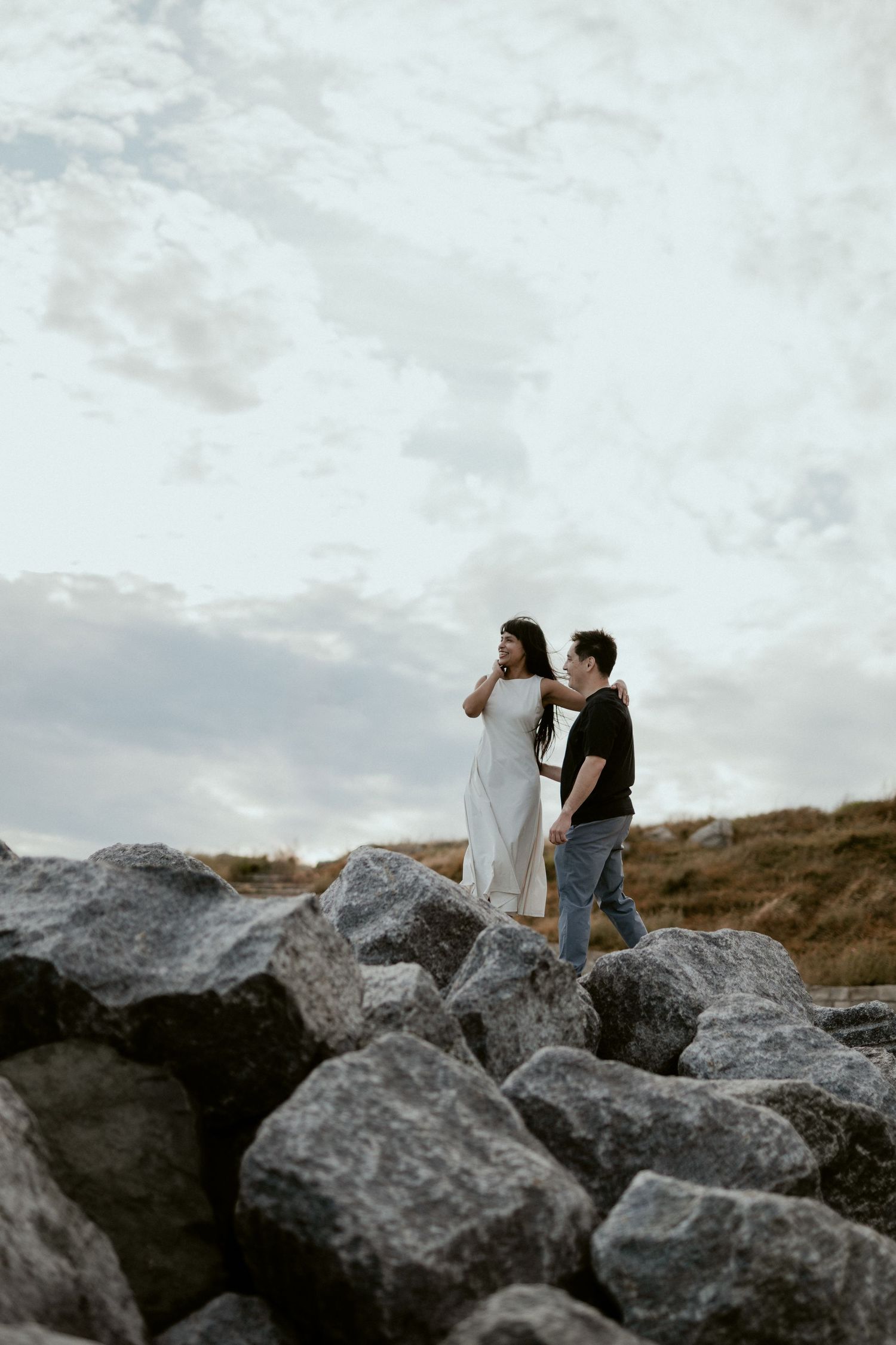 Couple in contrasting outfits posing dramatically on rocky coastal terrain against cloudy sky.