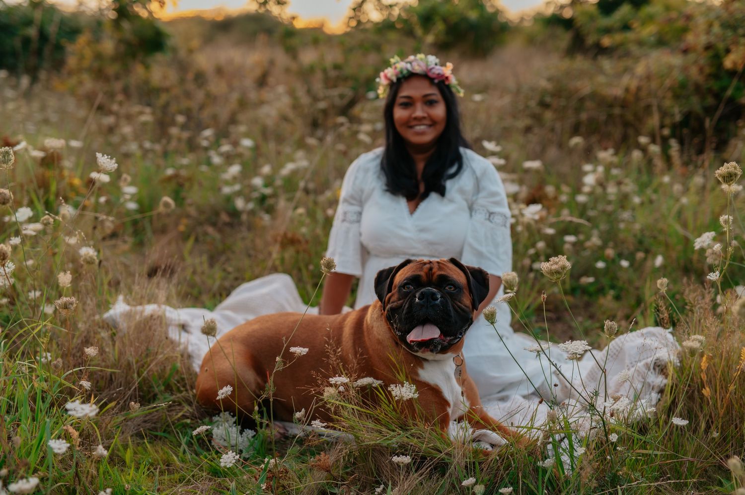 Woman in white dress poses with a brown dog in a field of white flowers.