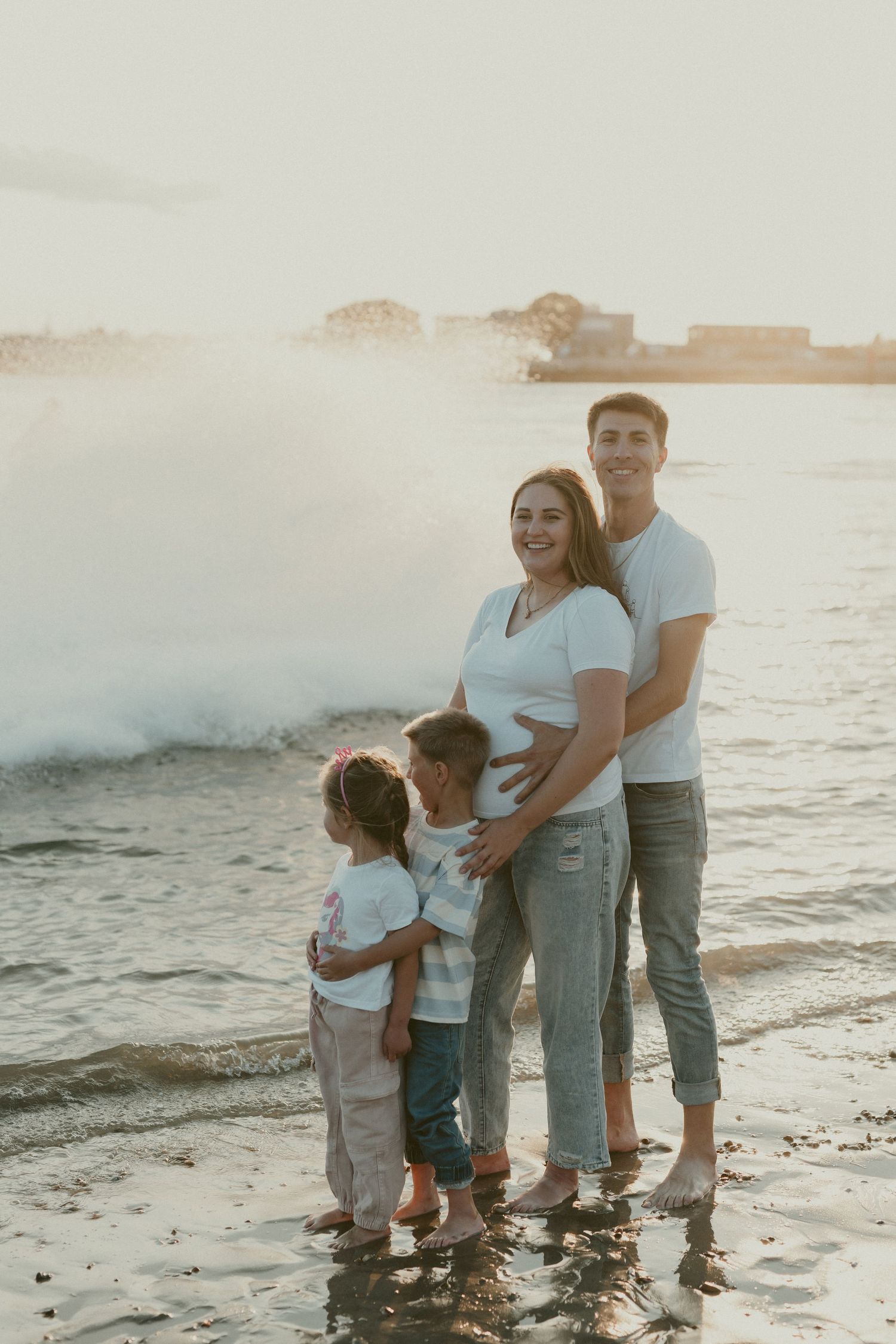 Family of four standing together on beach shoreline during sunset.