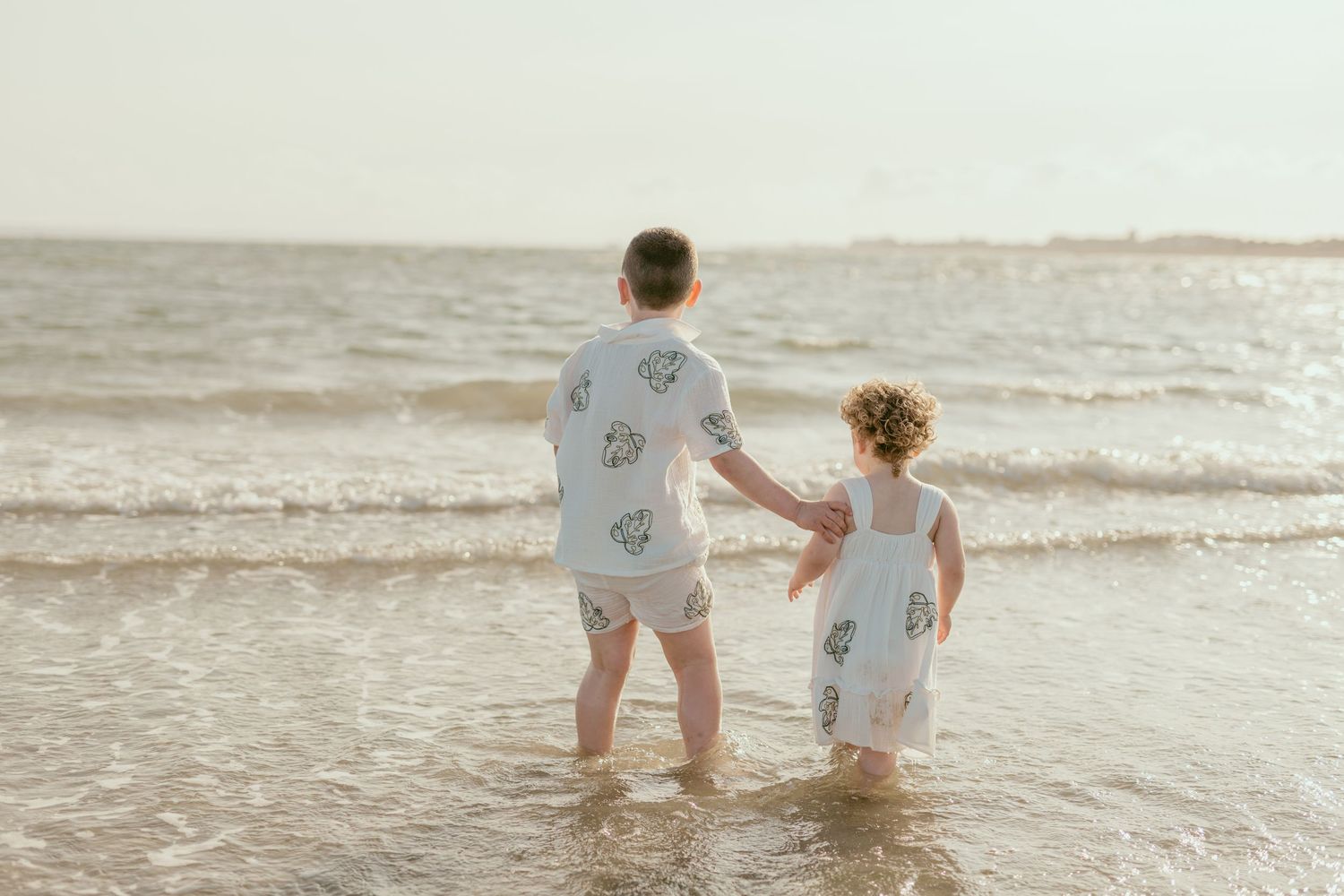 Two people in matching white outfits walk together in shallow ocean water.
