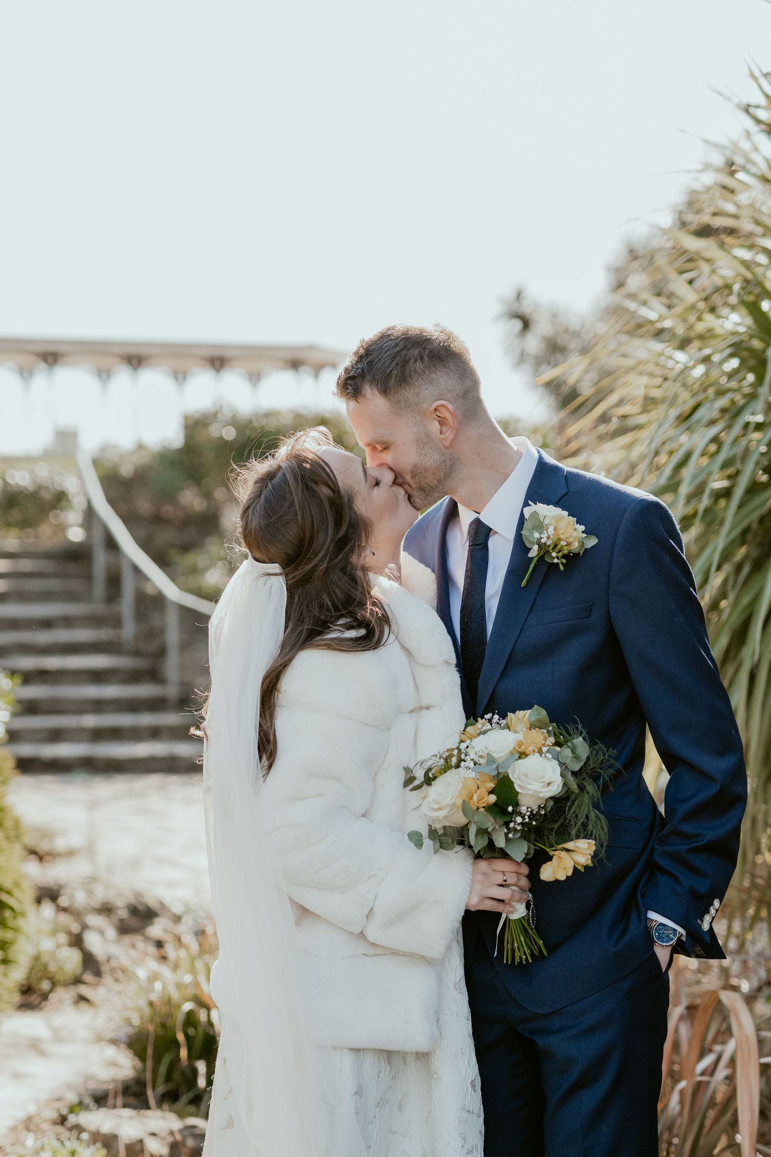 Wedding couple embraces outdoors against a backdrop of coastal grasses and natural scenery.