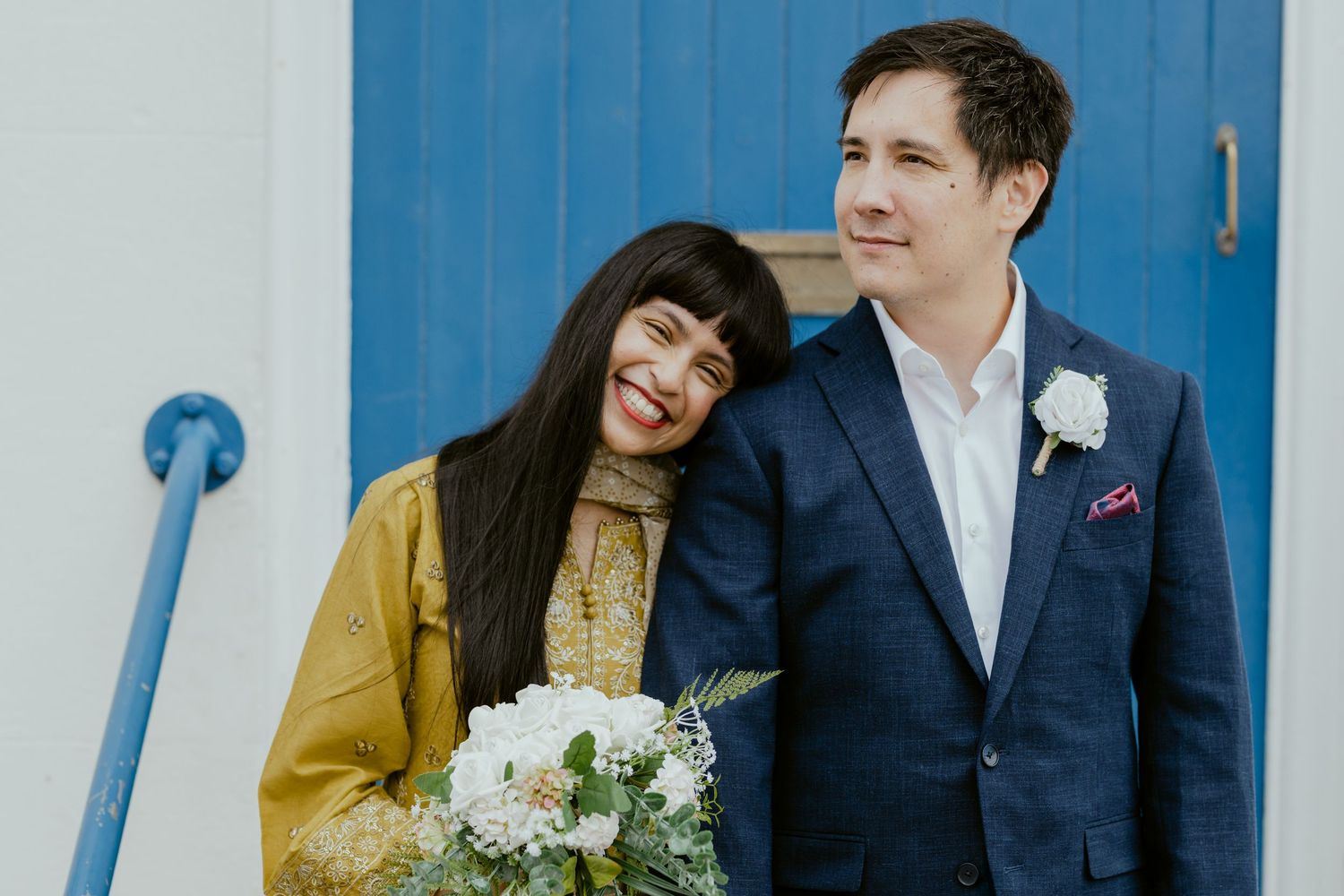 Couple in wedding attire posing against white wall with bright blue door.