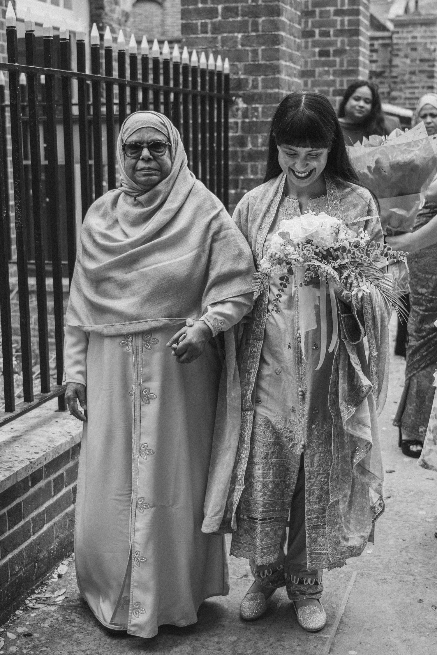 A black and white photo of two people walking together, one in traditional dress holding a floral bouquet.