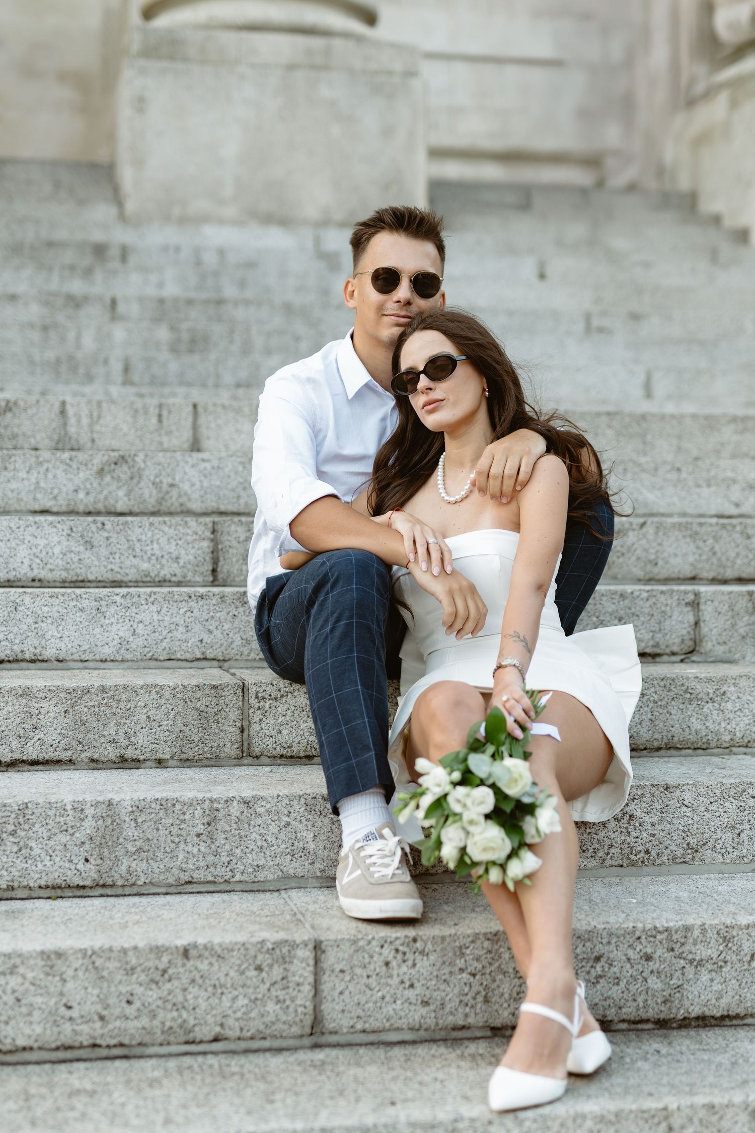 Couple relaxes on stone steps with white flowers during romantic outdoor photoshoot.