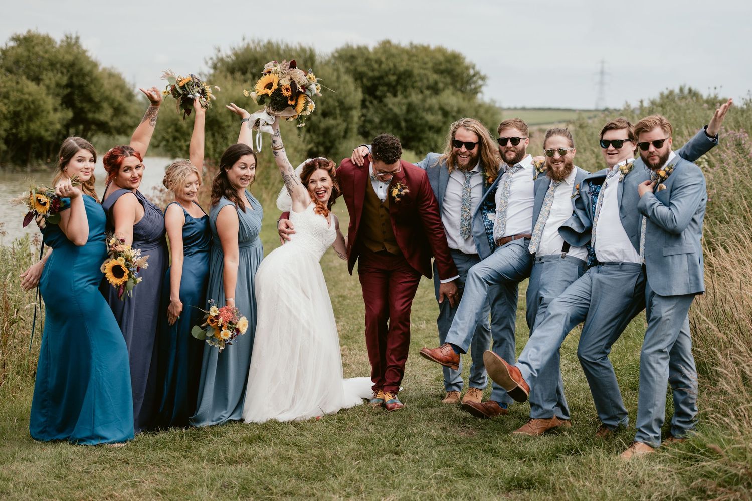 Wedding party in teal dresses and gray suits poses playfully on grassy field.