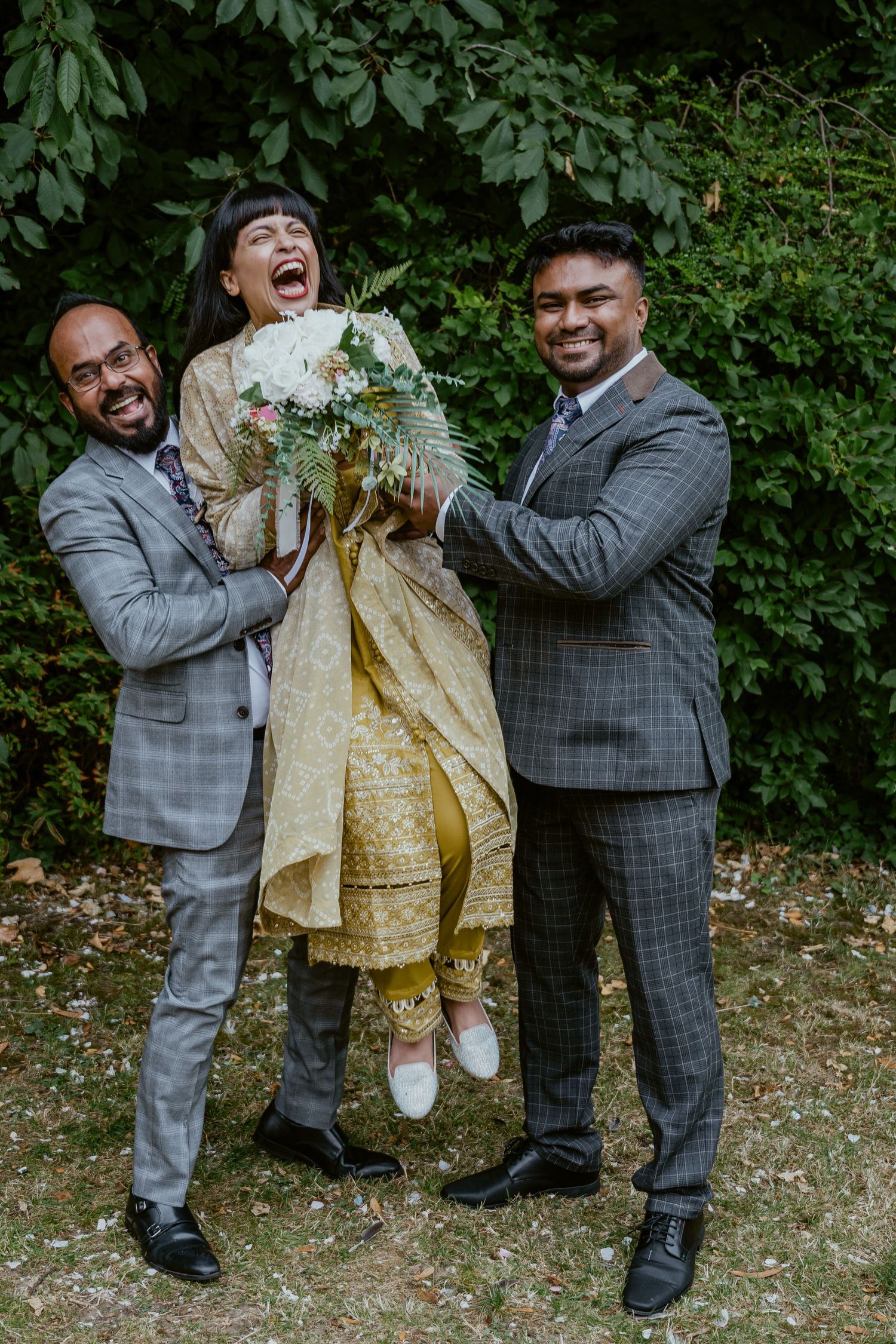 Three people share a joyful moment lifting someone in a gold outfit against green foliage.