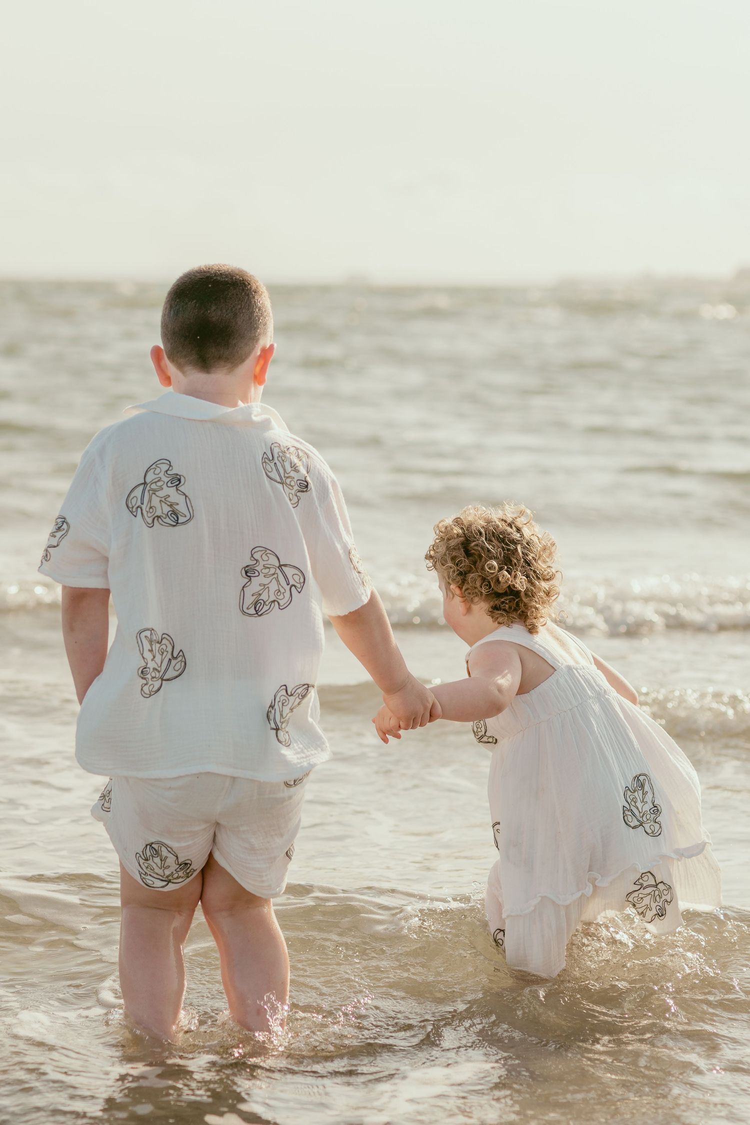 Parent and child holding hands while standing in ocean waves wearing matching outfits.