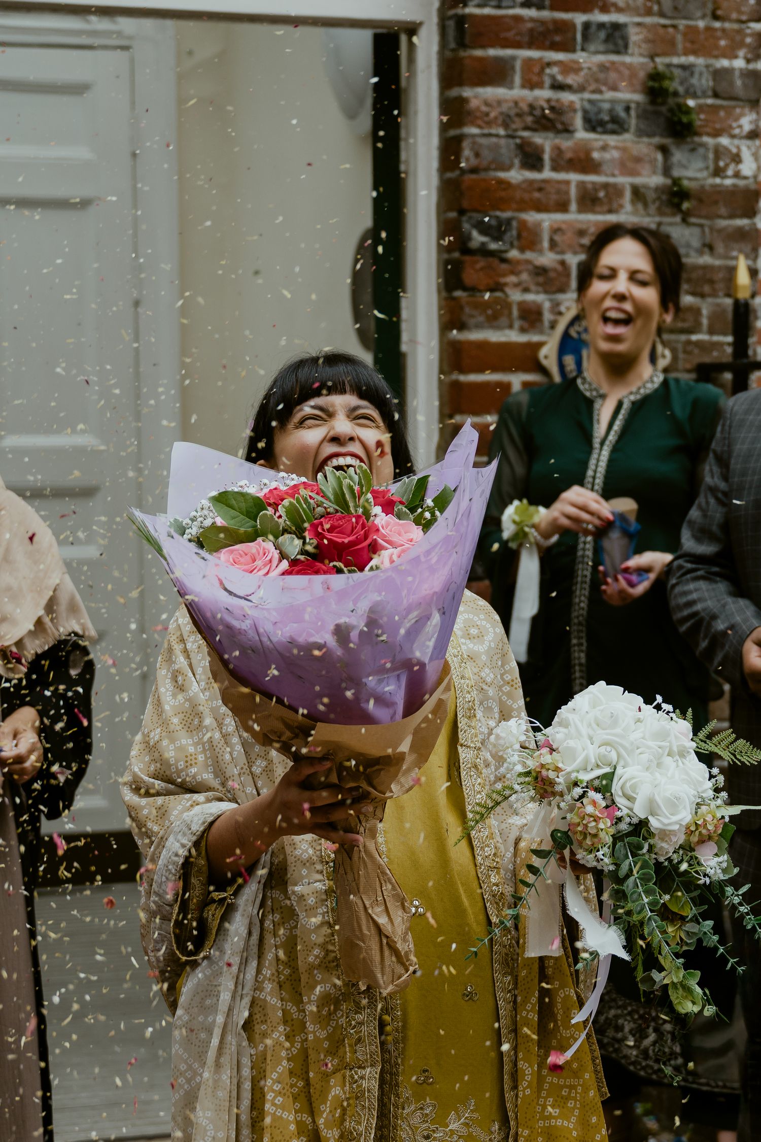 A joyful moment captured outside as confetti falls around someone holding a purple-wrapped flower bouquet.
