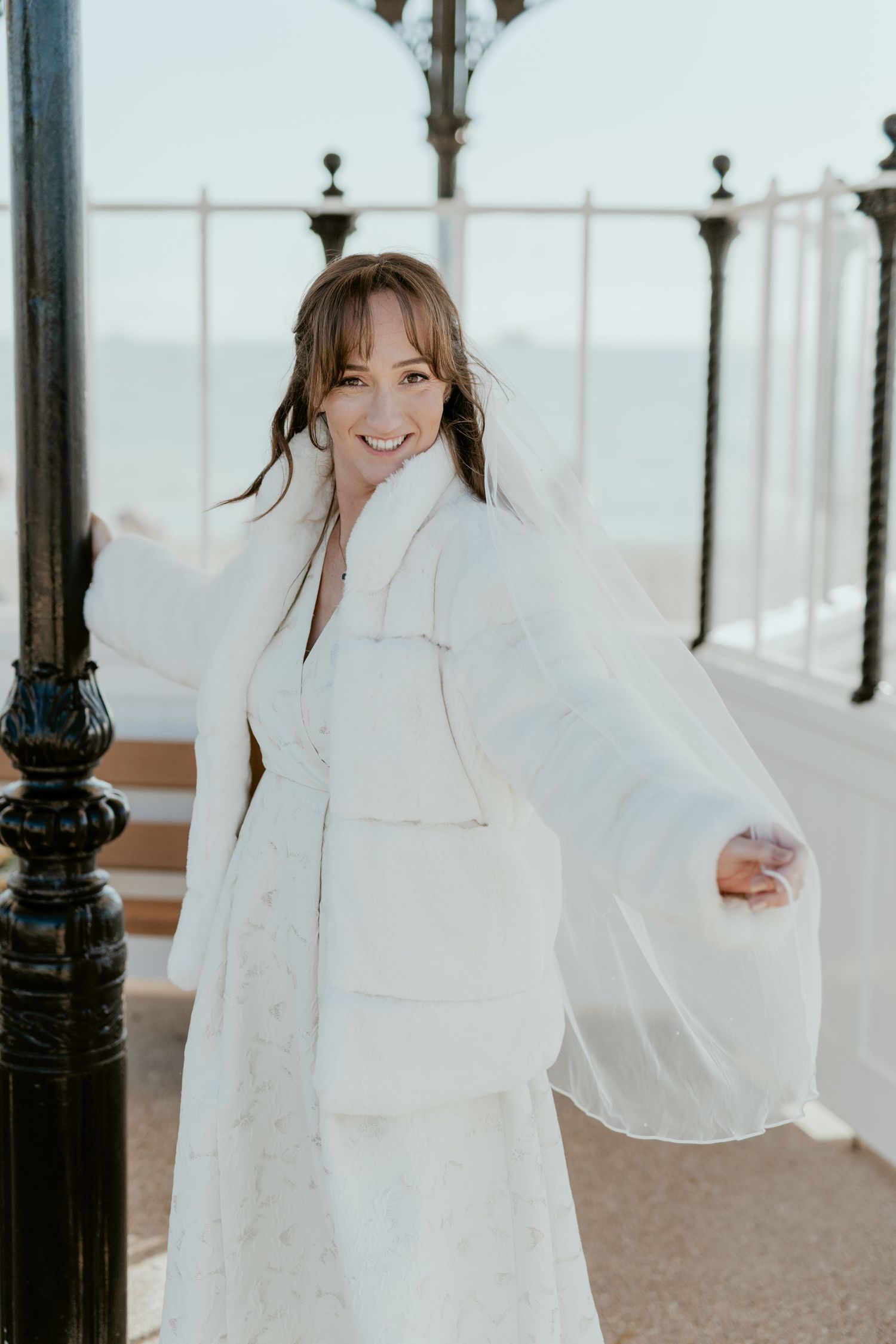 Bride in a flowing white dress twirls joyfully on a bright seaside promenade.